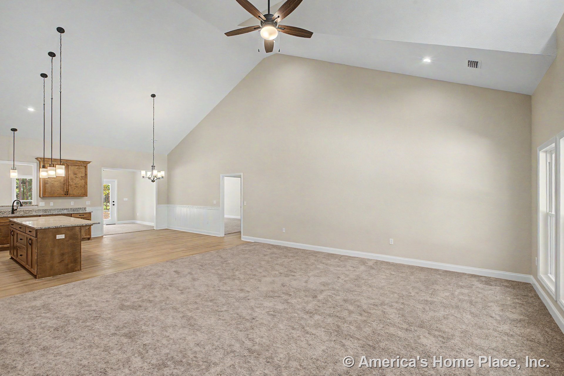 Vaulted ceiling with ceiling fan above spacious carpeted living area, open to kitchen featuring granite island, wood cabinetry, pendant lighting, wide windows with white trim, and