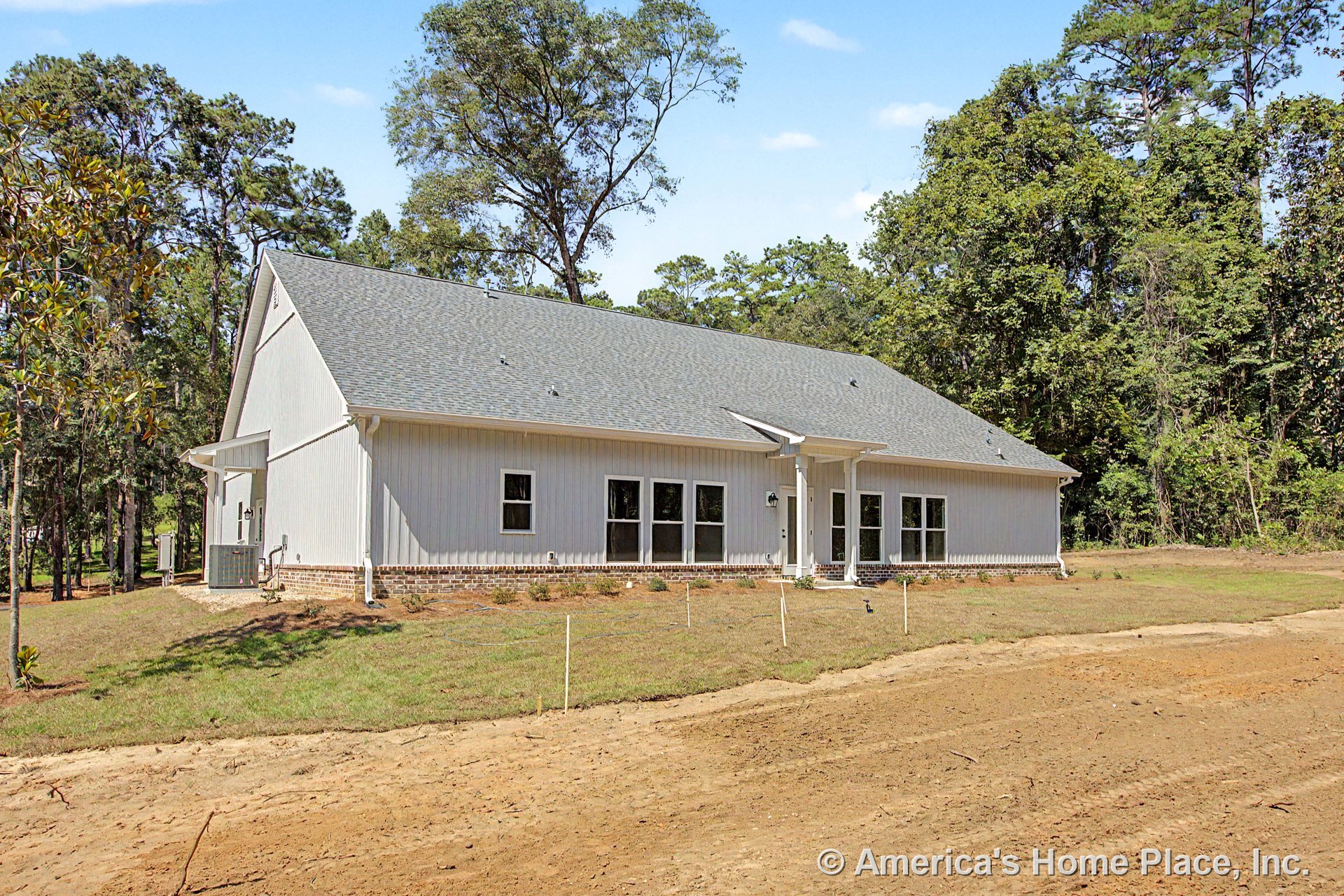 Gray gabled shingle roof, vertical board siding, brick foundation trim, covered front porch with white exterior doors, multiple rectangular windows, outdoor lighting fixtures.