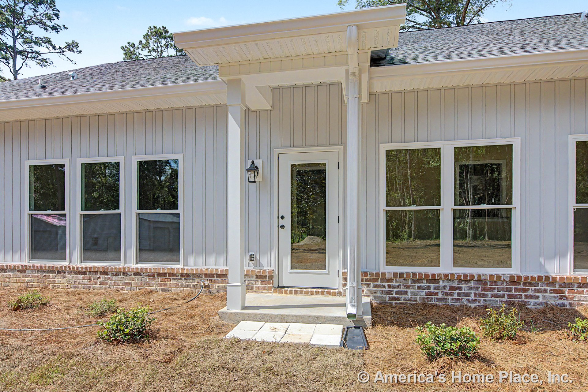 Vertical panel siding with brick foundation trim, covered porch entry supported by columns, glass exterior door, multiple large windows, outdoor wall lantern, roof overhang