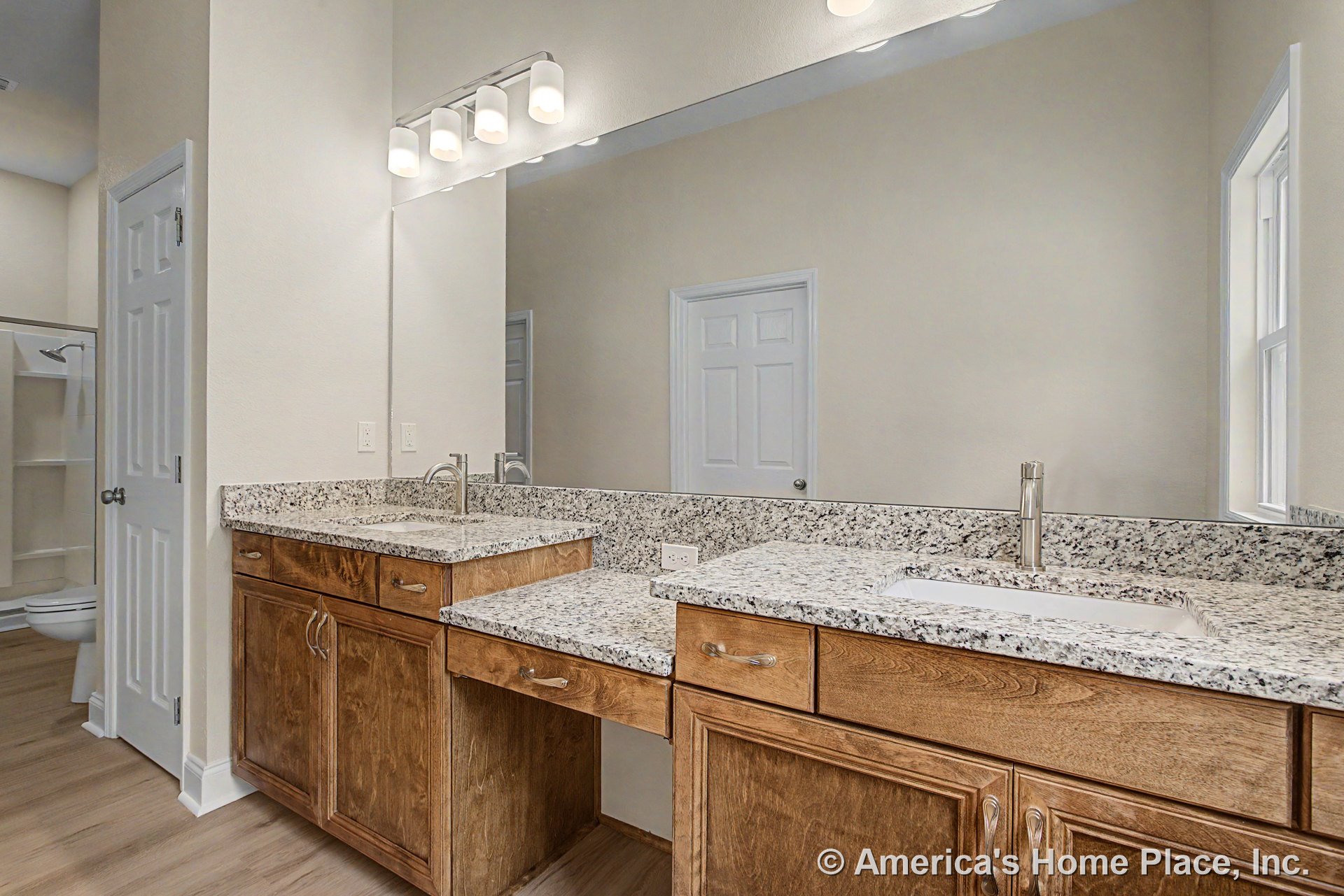 Double vanity with granite countertops, wood cabinetry, two sinks, large wall mirror, built-in vanity lighting, adjacent walk-in shower, window, and door with trim.