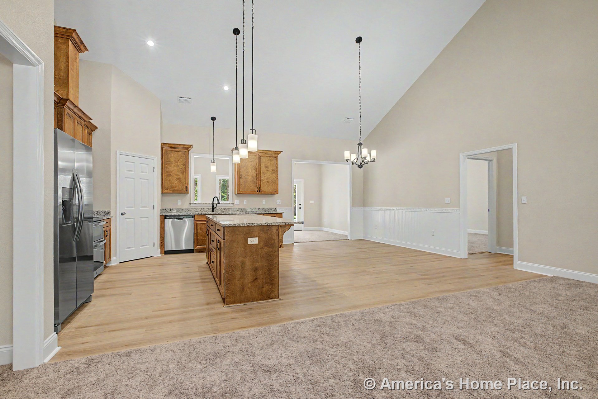 Open kitchen with wood cabinetry, granite countertops, stainless steel refrigerator, and kitchen island, adjoining a vaulted ceiling dining area with wainscoting trim, pendant and