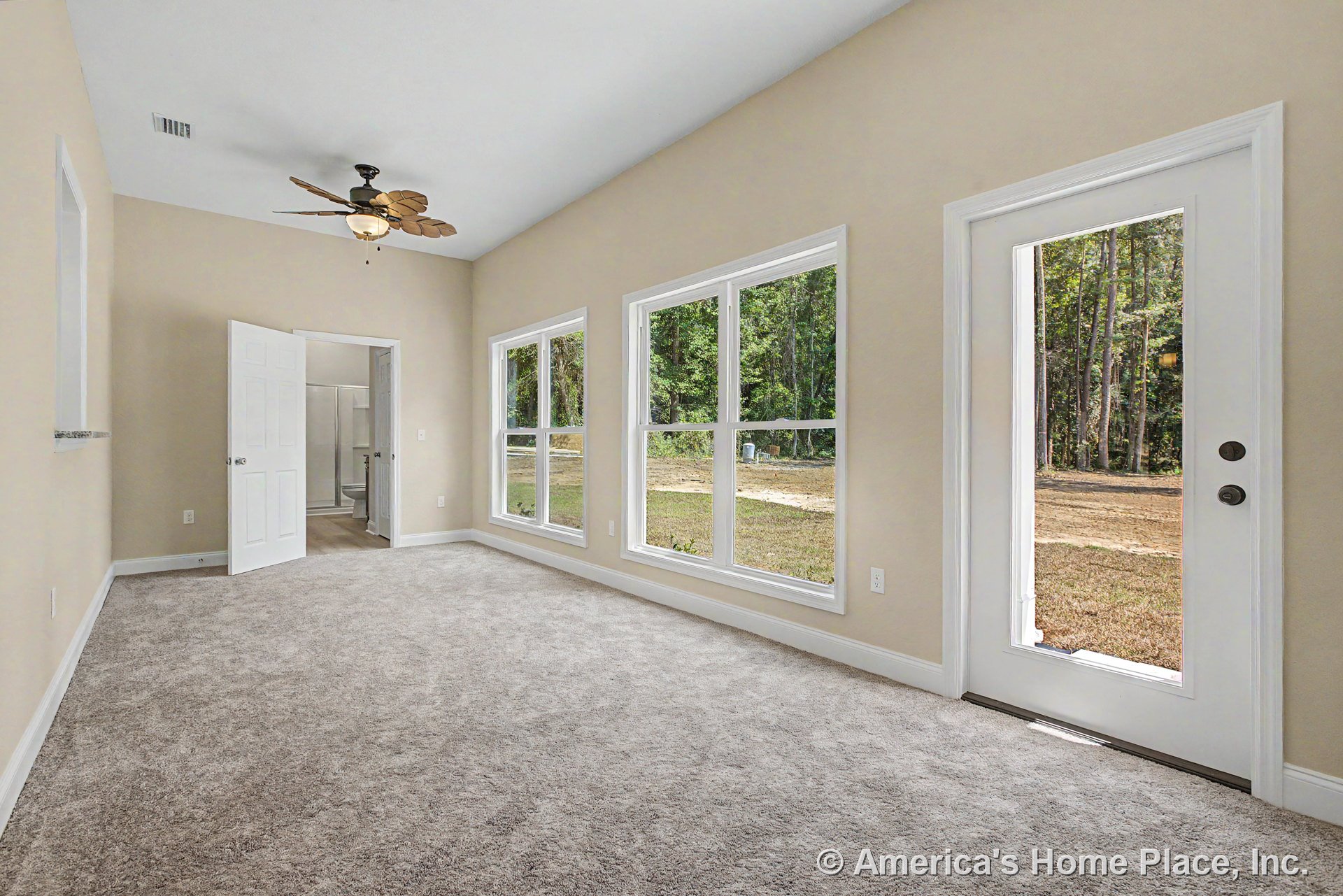 Carpeted living room with three large windows, glass exterior door, ceiling fan with light fixture, white trim and baseboards, and neutral wall color.