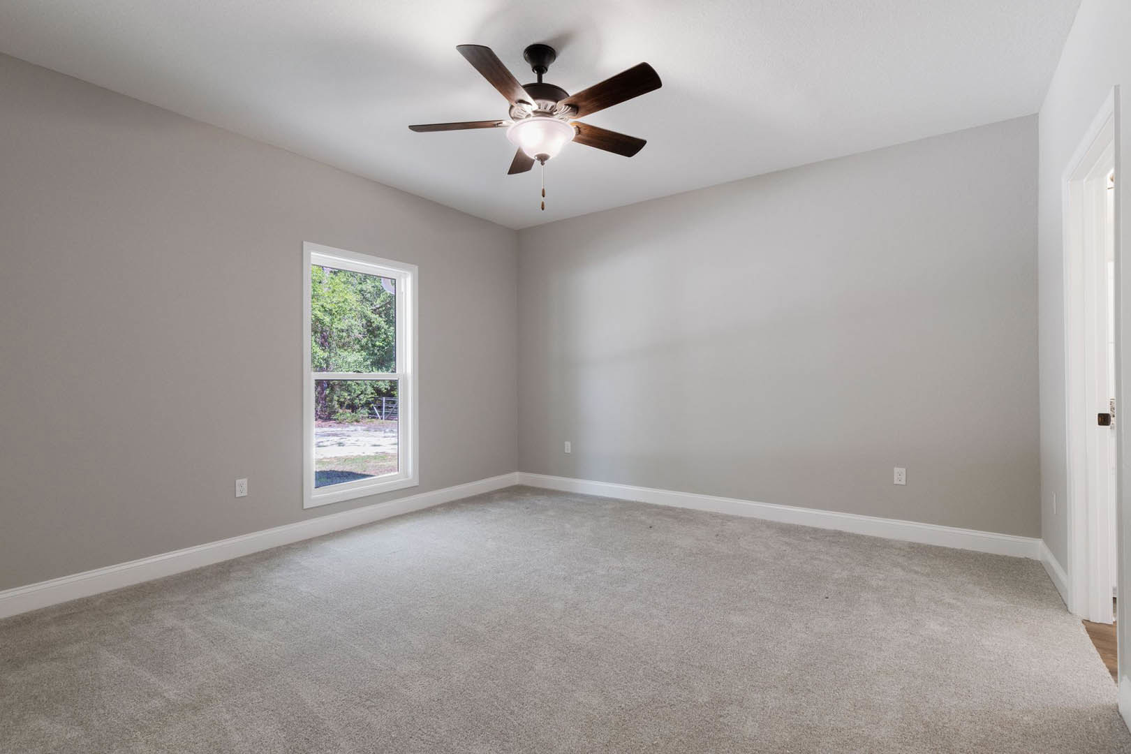 Carpeted room with white walls, ceiling fan with light fixture, large window overlooking trees, simple crown molding along ceiling.