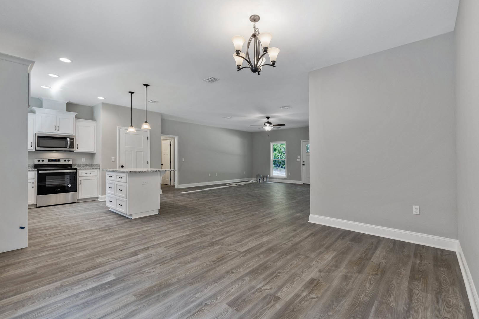 Spacious kitchen with wood flooring, five-light chandelier, white cabinetry, stove beneath a window overlooking trees, plaster walls, and a person standing near the window.