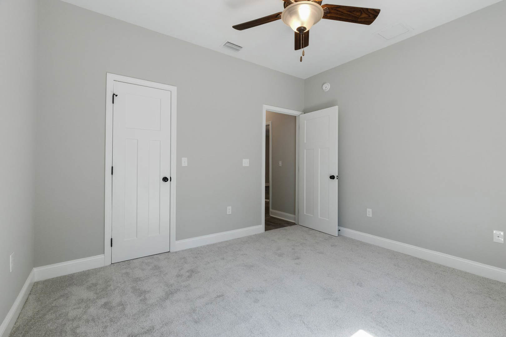 Carpeted room with white walls, ceiling fan with light fixture, two white doors with black knobs, and visible light switch.