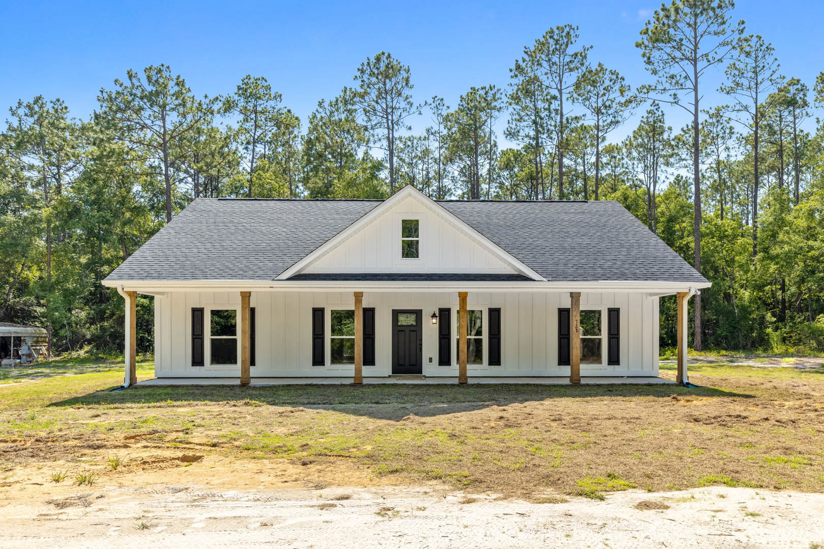 White house with black shutters and black door, rectangular window, grassy yard with patches of dirt, group of trees and blue sky in background, smaller white house visible behind