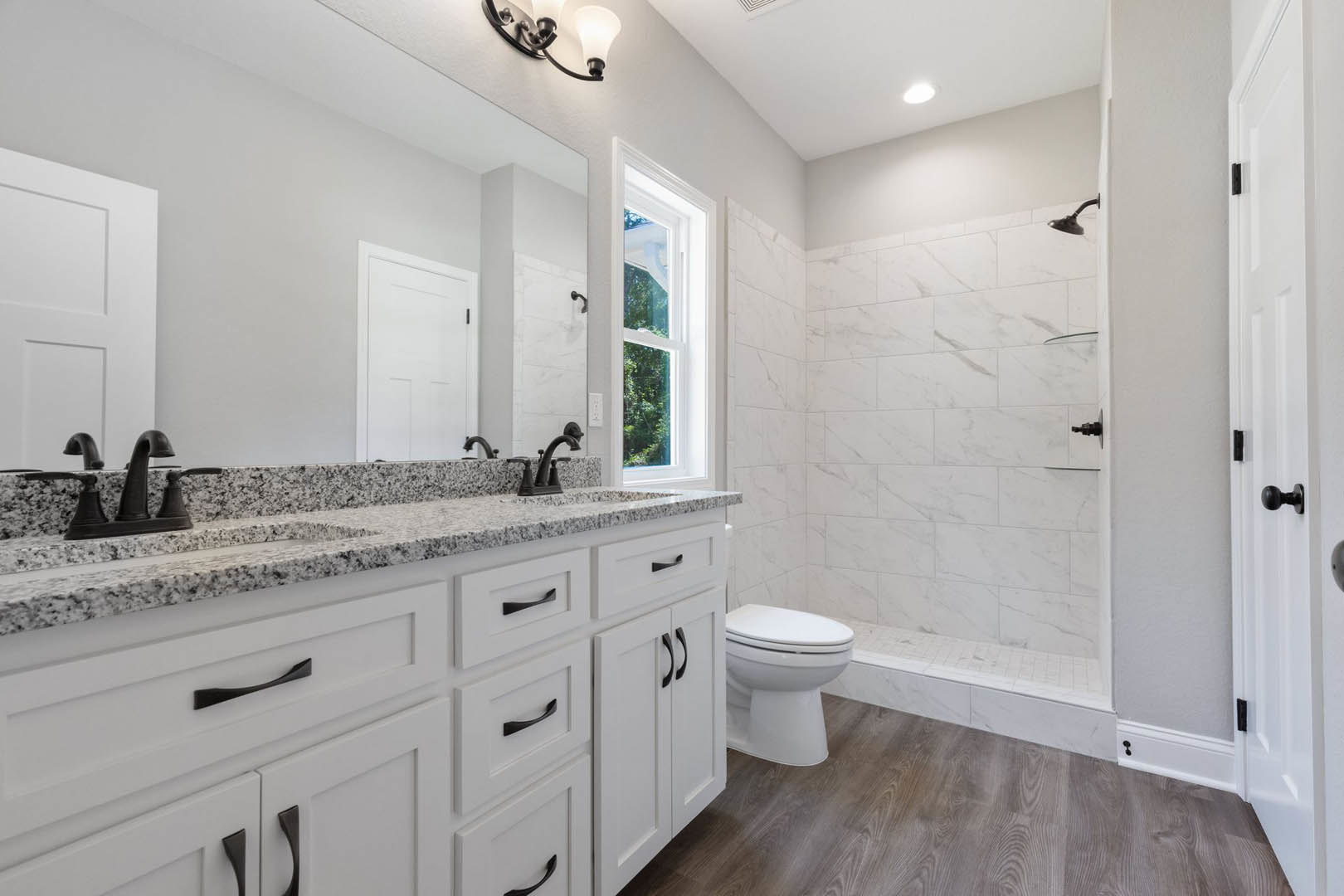 Bathroom with marble countertop, undermount sink, chrome faucet, glass-enclosed shower, white toilet, white framed window, white door with black hinges, wall sconce above vanity