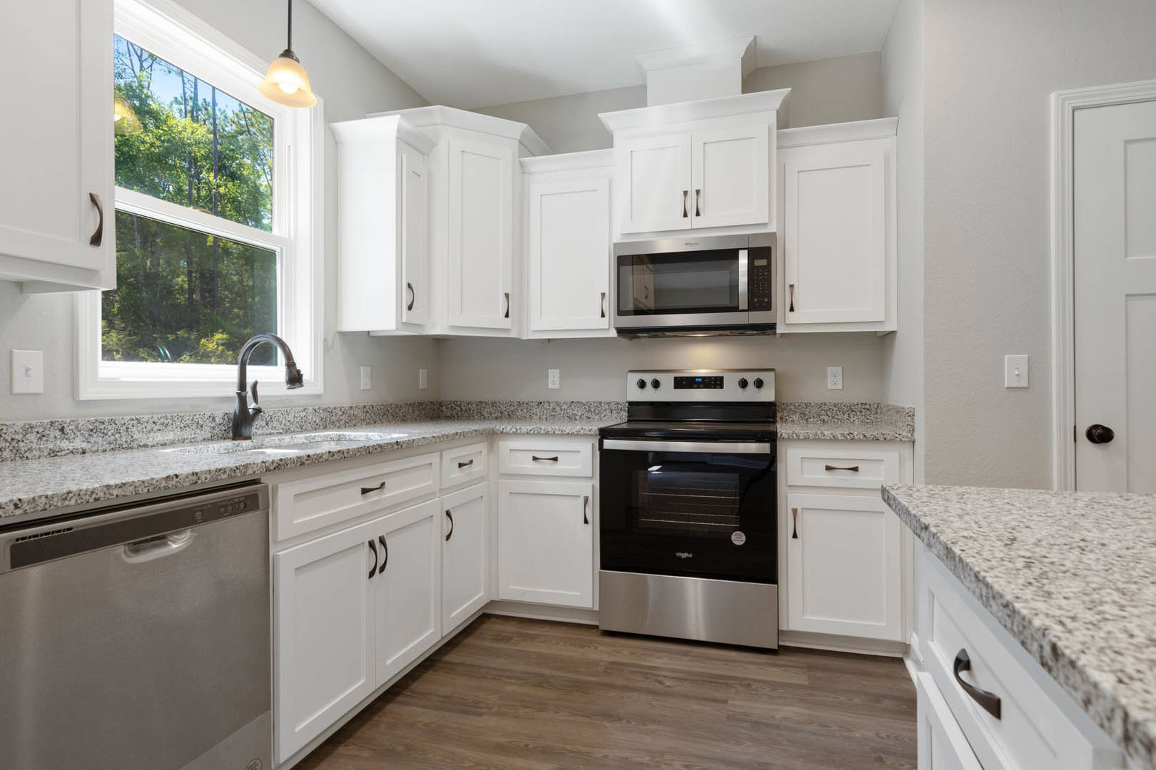White kitchen cabinets and granite countertops with a black and silver oven, glass door microwave, close-up dishwasher, ceiling light fixture, and window overlooking trees.