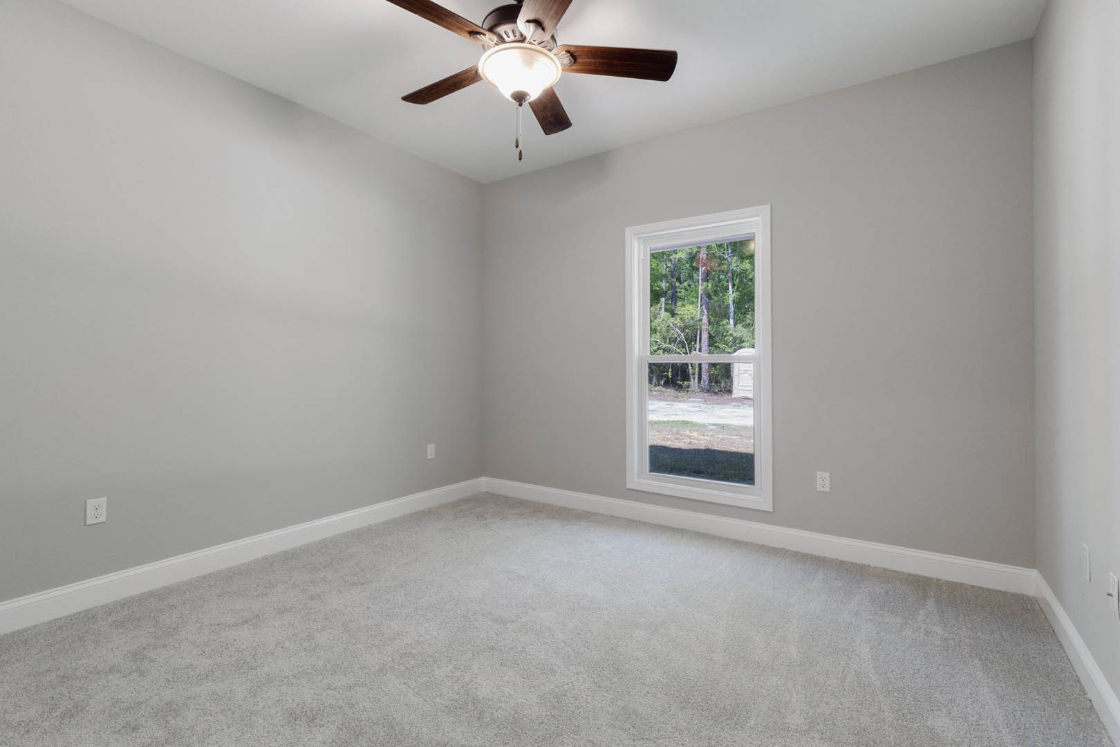 Neutral-toned room with white carpet, ceiling fan featuring light fixture, large window overlooking leafy trees, plaster walls, and crown molding.