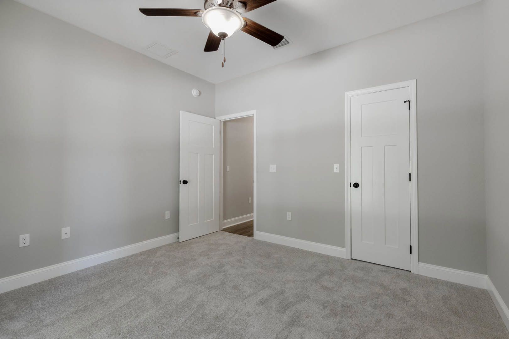 Bedroom with light gray carpet, white walls, ceiling fan with integrated light, two white doors featuring black hardware, and visible light switch near entry.