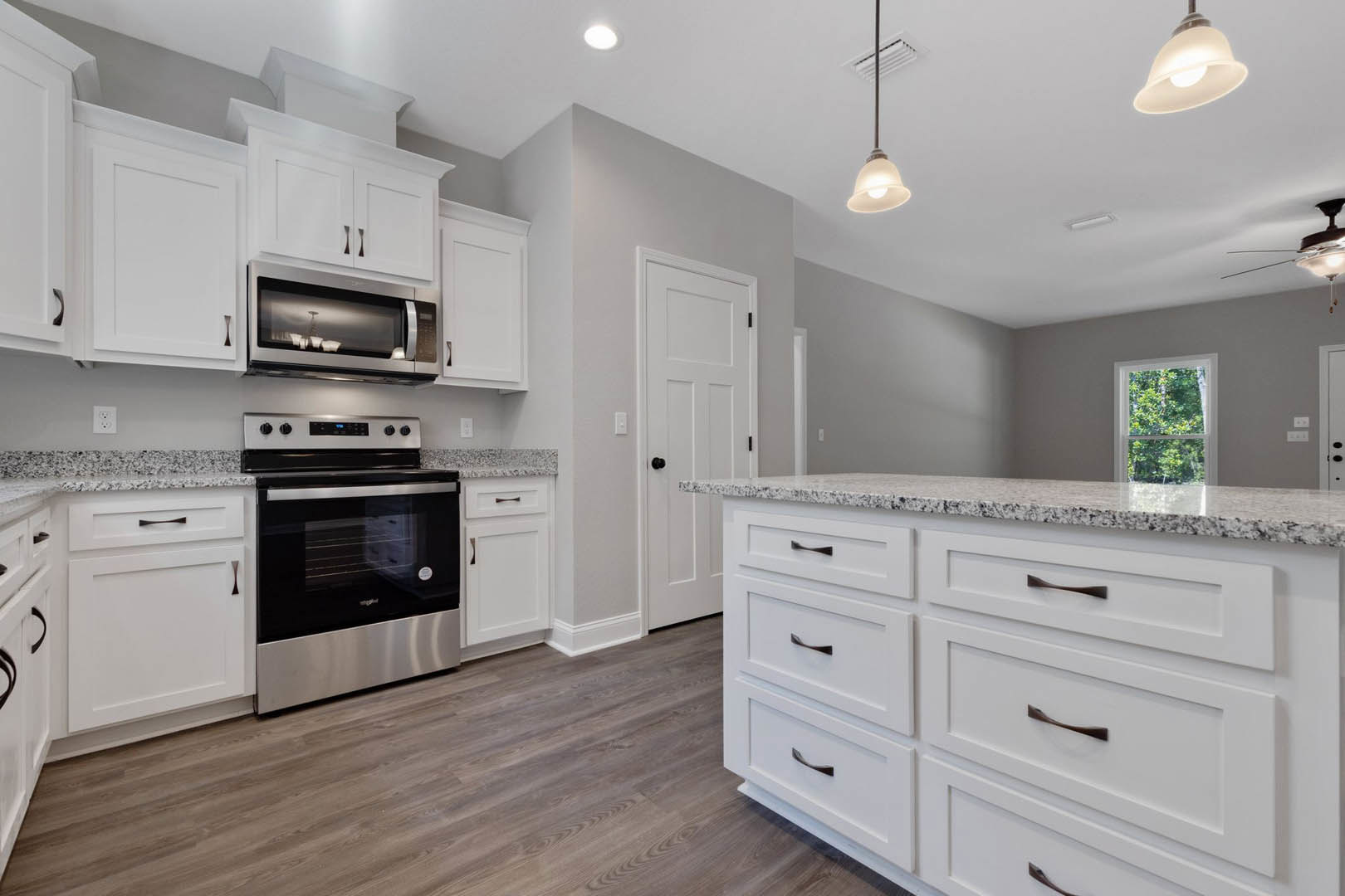 White kitchen with granite countertops, stainless steel oven and microwave, drawers beneath counter, pendant light fixture, window overlooking trees.