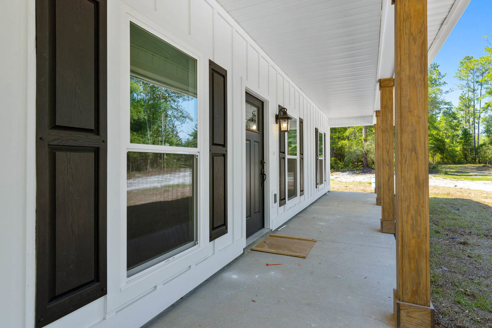 White siding house with black shutters, dark wood front door, porch with wooden flooring, multiple windows, shaded by nearby trees