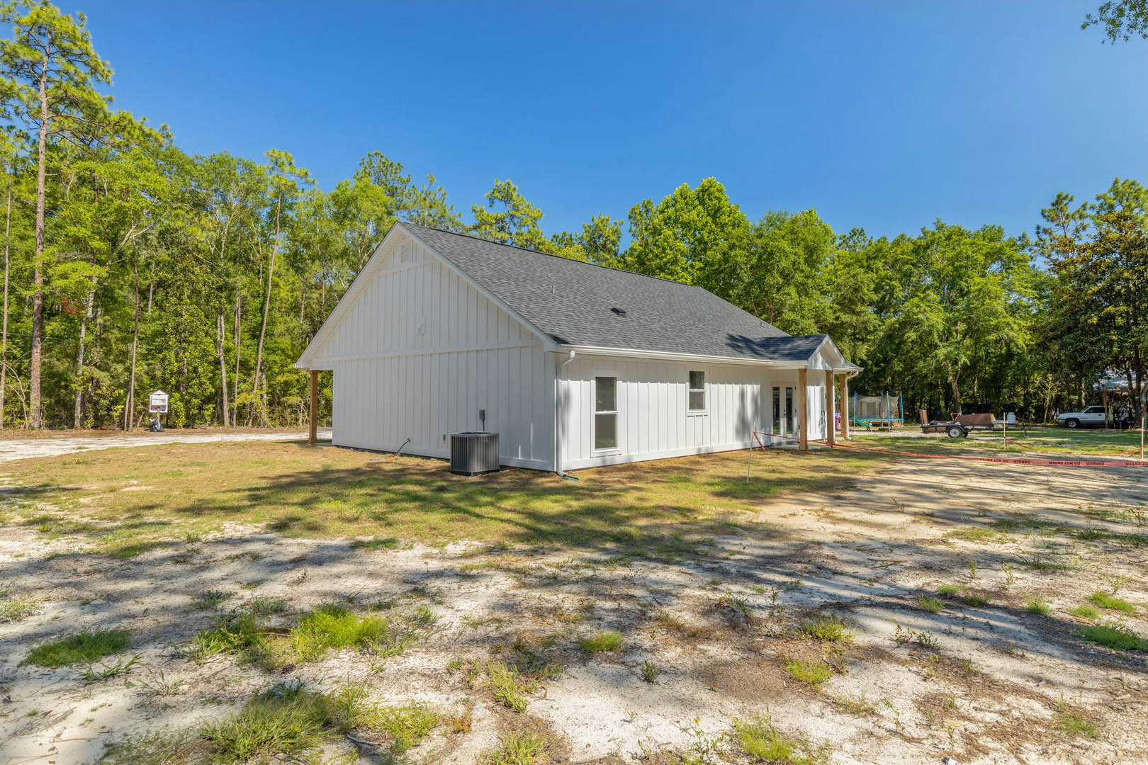 White farmhouse with gabled roof, grassy lawn, metal HVAC unit with red tag, surrounded by tall trees and forest backdrop