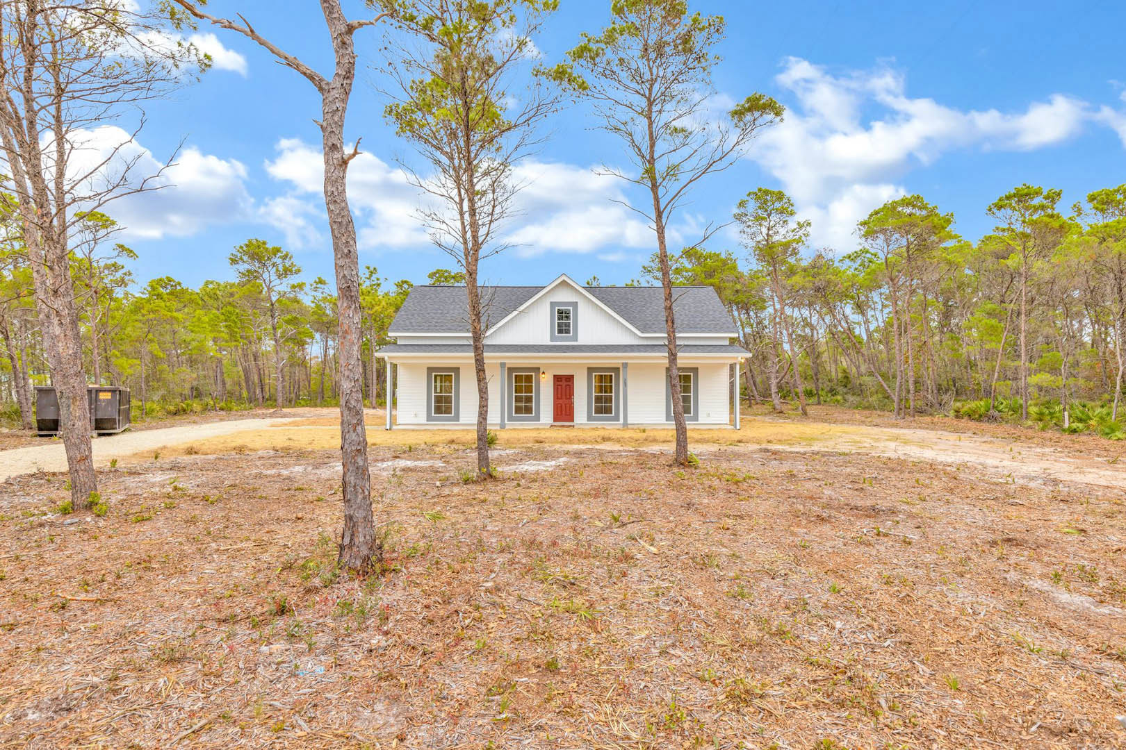 Two-story custom home with white siding, red front door, and large windows, surrounded by mature trees and green lawn under a partly cloudy sky