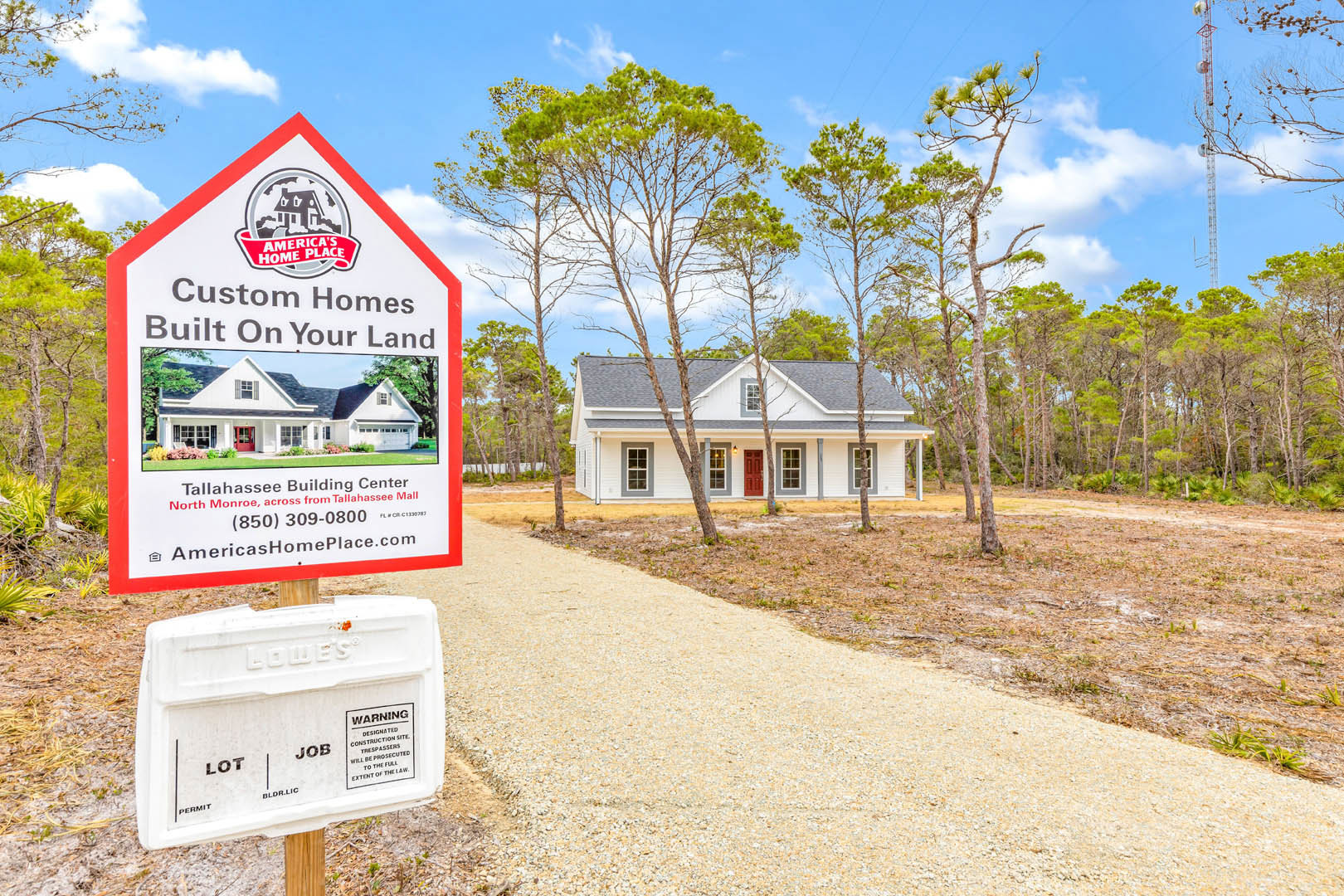 Wooden post with stacked signs beside a house featuring a red door, gravel driveway, and surrounding trees under a cloudy sky