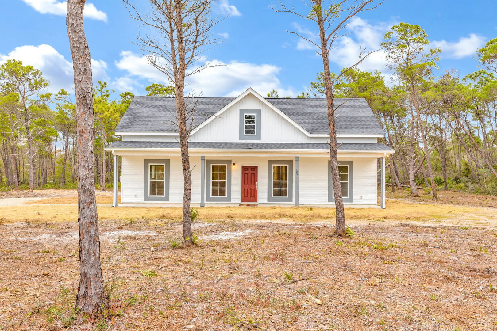 White-walled home with red front door, surrounded by mature trees and grassy lawn, white-framed windows visible on exterior facade