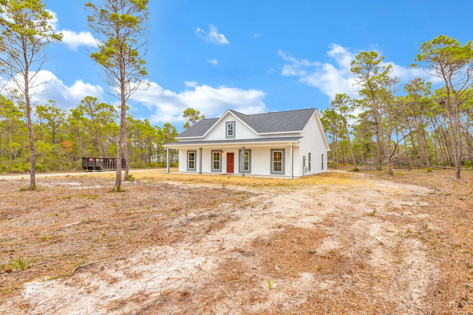 Two-story custom home with gray siding, red front door, surrounded by mature trees and patchy grass, cloudy sky overhead