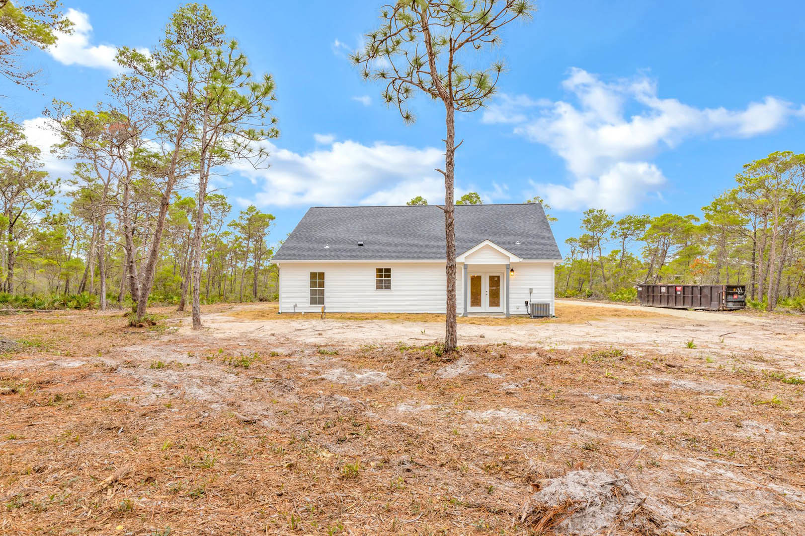 White farmhouse surrounded by mature trees, brown planter with red sign near dirt patch, tall tree in foreground, blue sky with scattered clouds