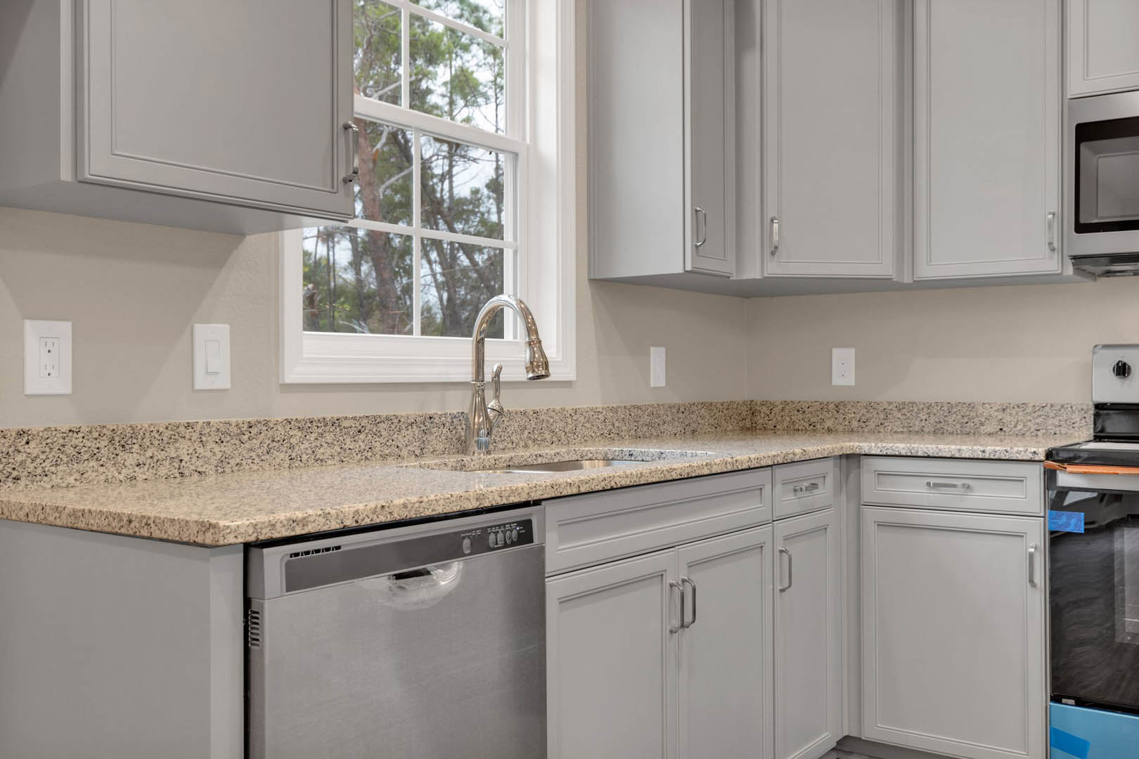 Marble countertop with undermount sink, chrome faucet, white cabinetry, built-in dishwasher, large window framing leafy trees, white cabinet handles, black-bordered appliance