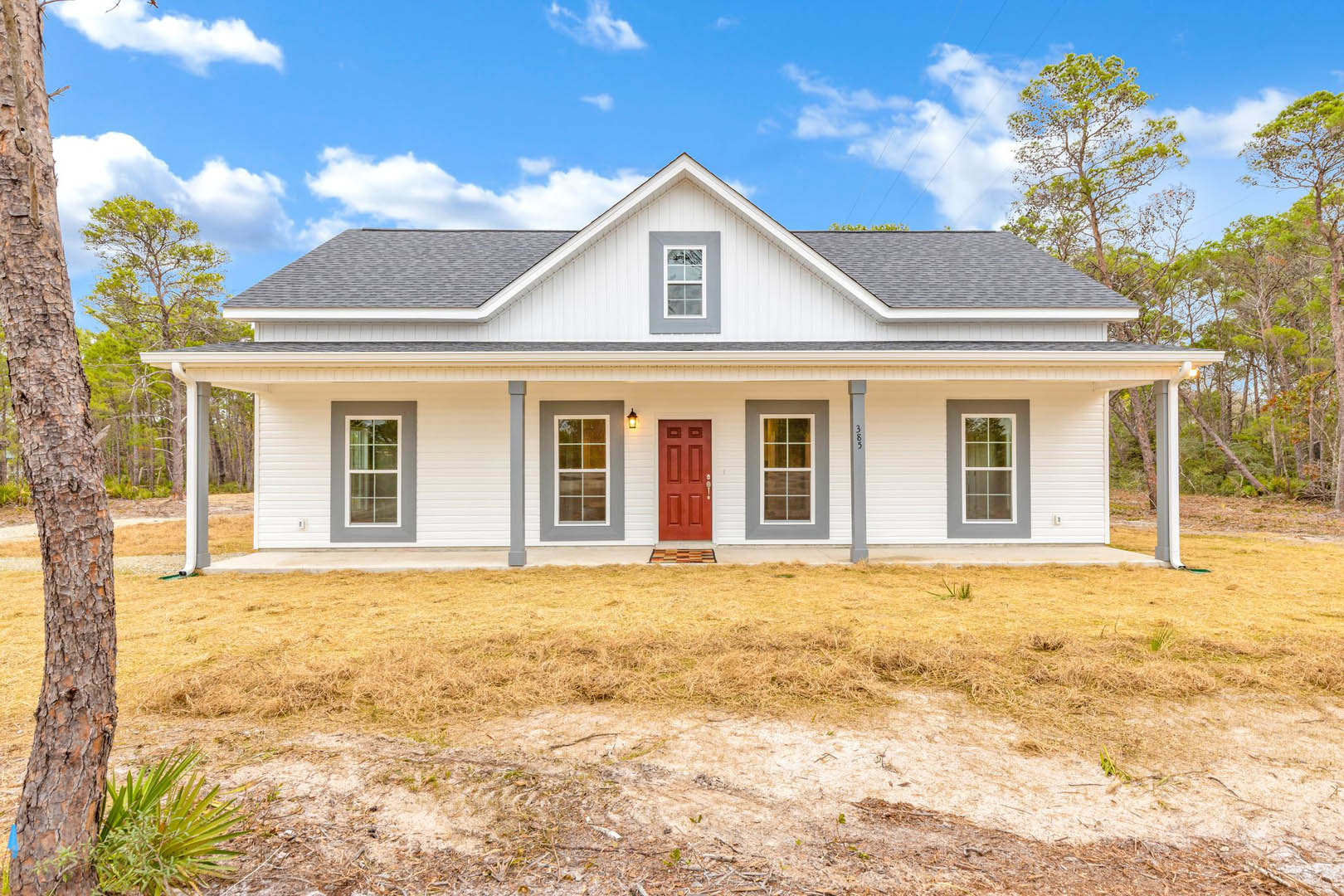 White siding exterior with a bold red front door, white-framed windows, covered porch, and landscaped greenery under a partly cloudy sky