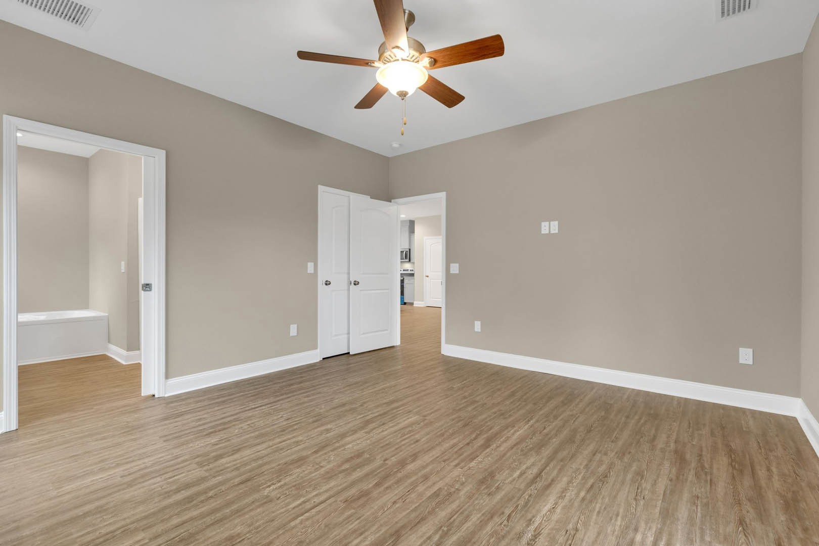 Ceiling fan with wooden blades and light fixture above wood flooring, white walls with trim, white door with silver knobs, partial view of bathroom with white bathtub