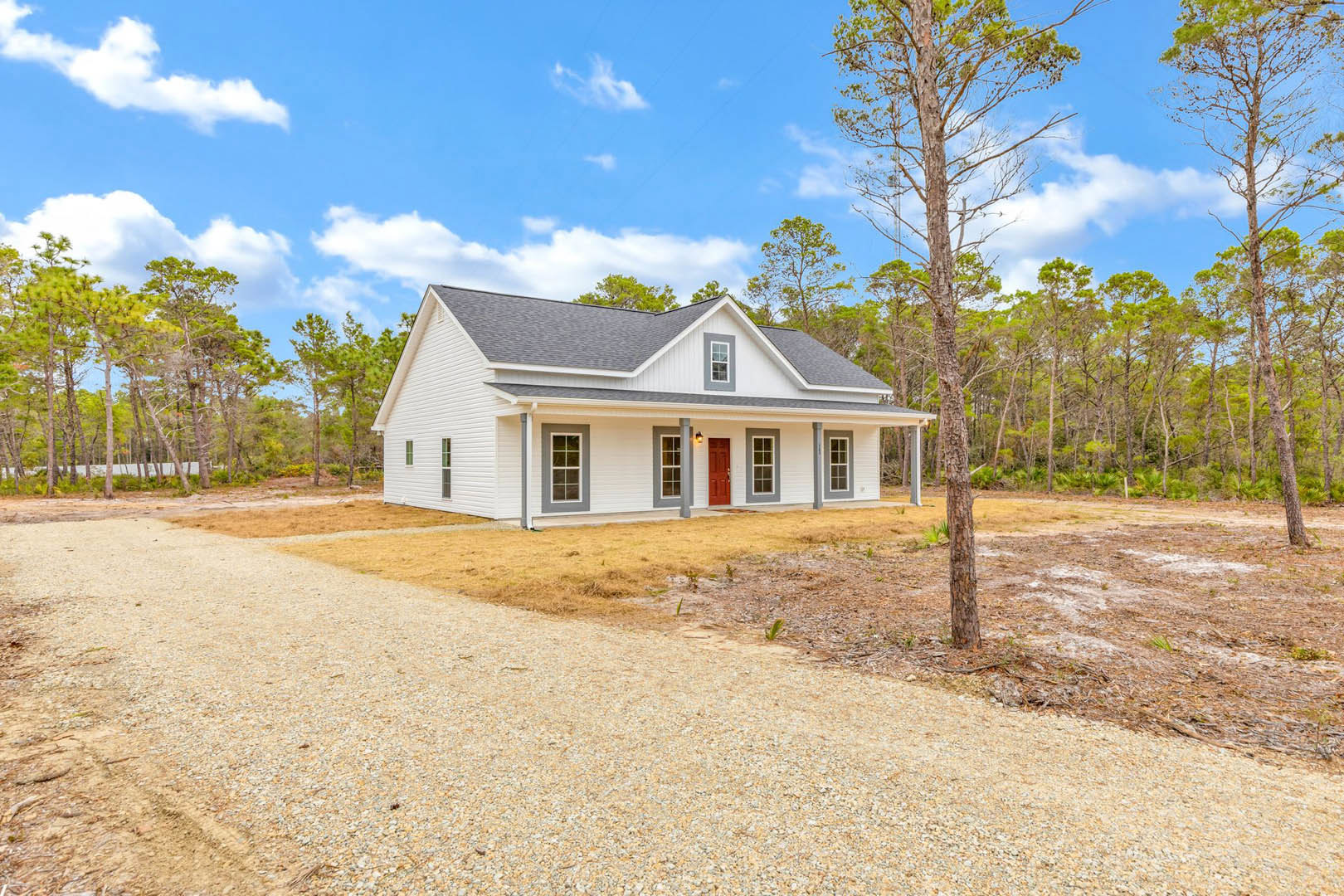 White house with gray trim, gravel driveway, mature trees in front and background, white-framed windows, grassy yard under partly cloudy sky