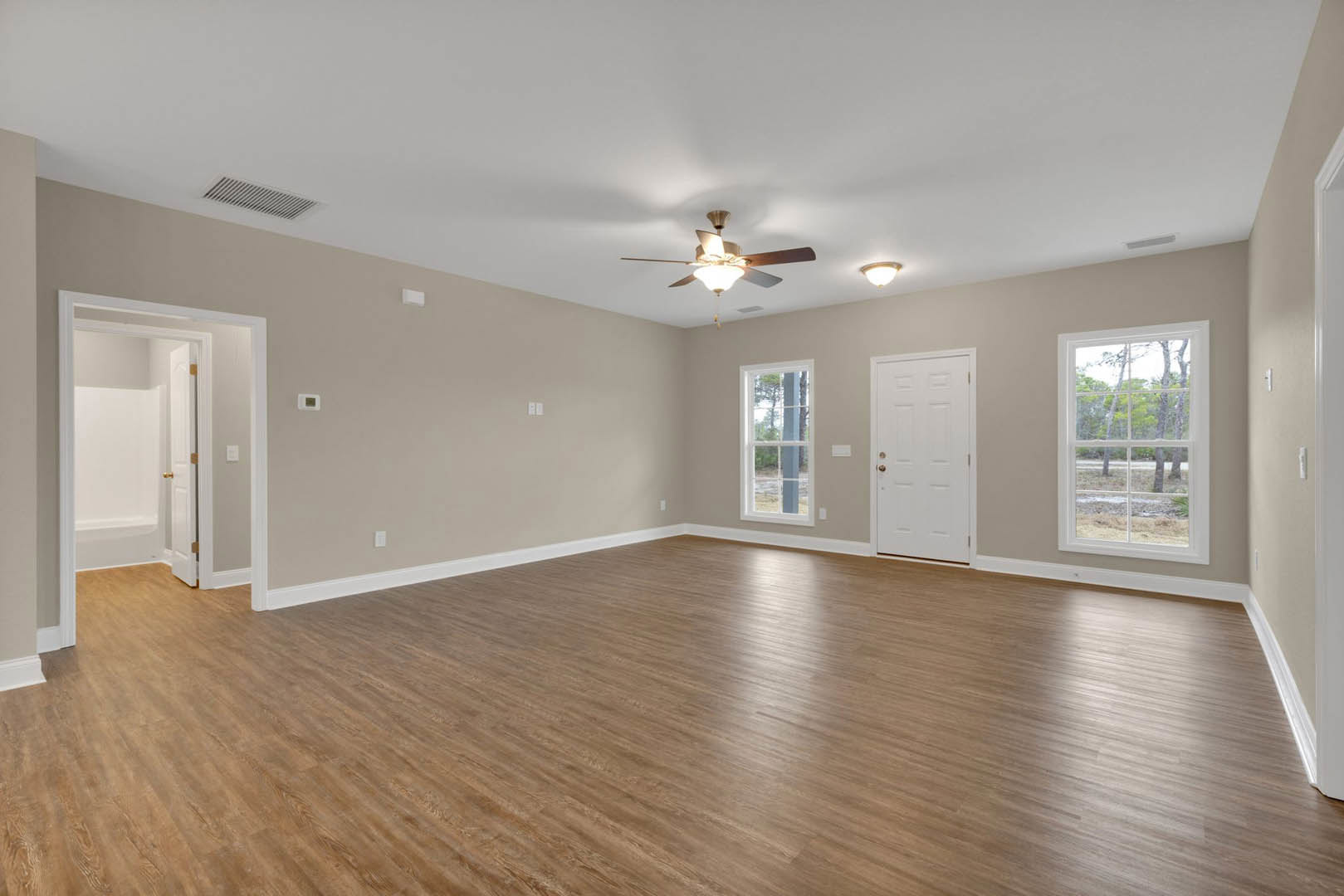 Wood flooring room with white walls, ceiling fan with light, white door featuring gold doorknob, and window framed in white showing trees outside