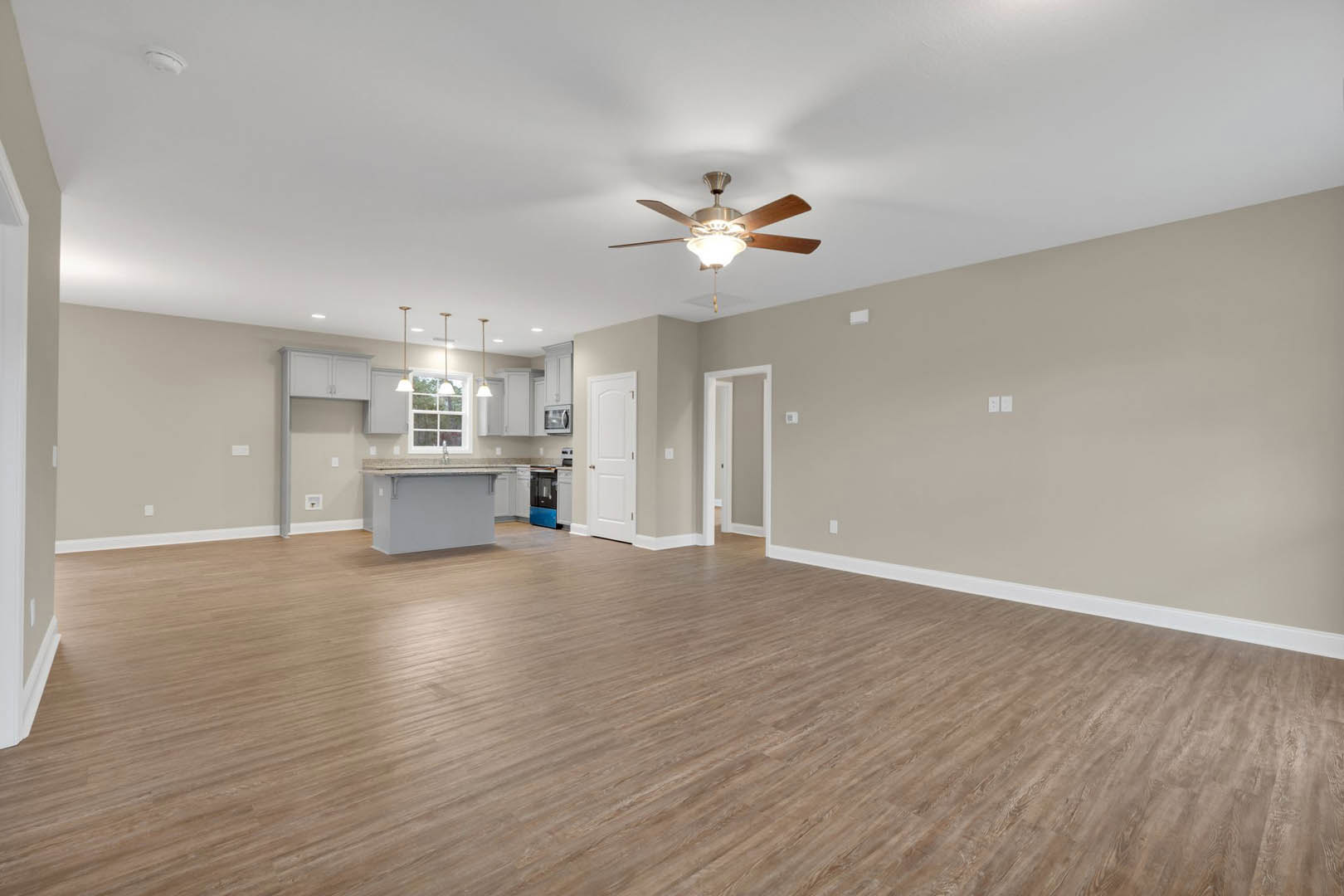 Open-concept room with wood laminate flooring, white walls, ceiling fan with light fixture, visible kitchen area, and white door with silver hardware