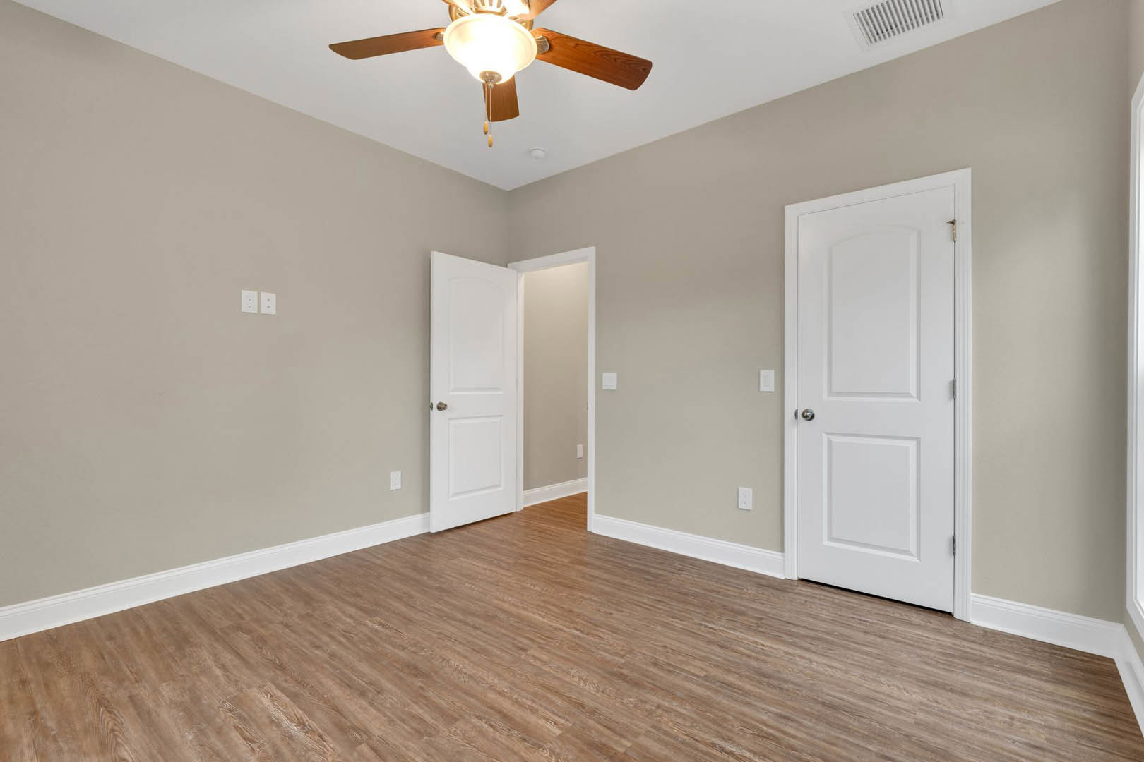 Room with wood flooring and white trim, two white doors with silver and gold knobs, ceiling fan with light fixture on white ceiling.