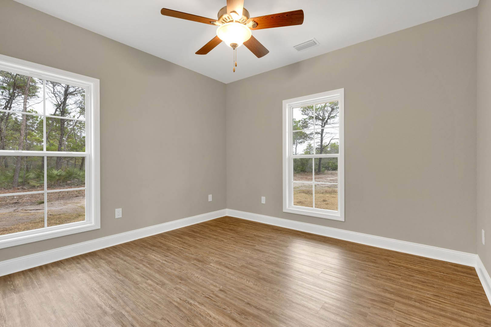 Ceiling fan with light fixture above wood flooring, white baseboards, and large windows showing green trees outside