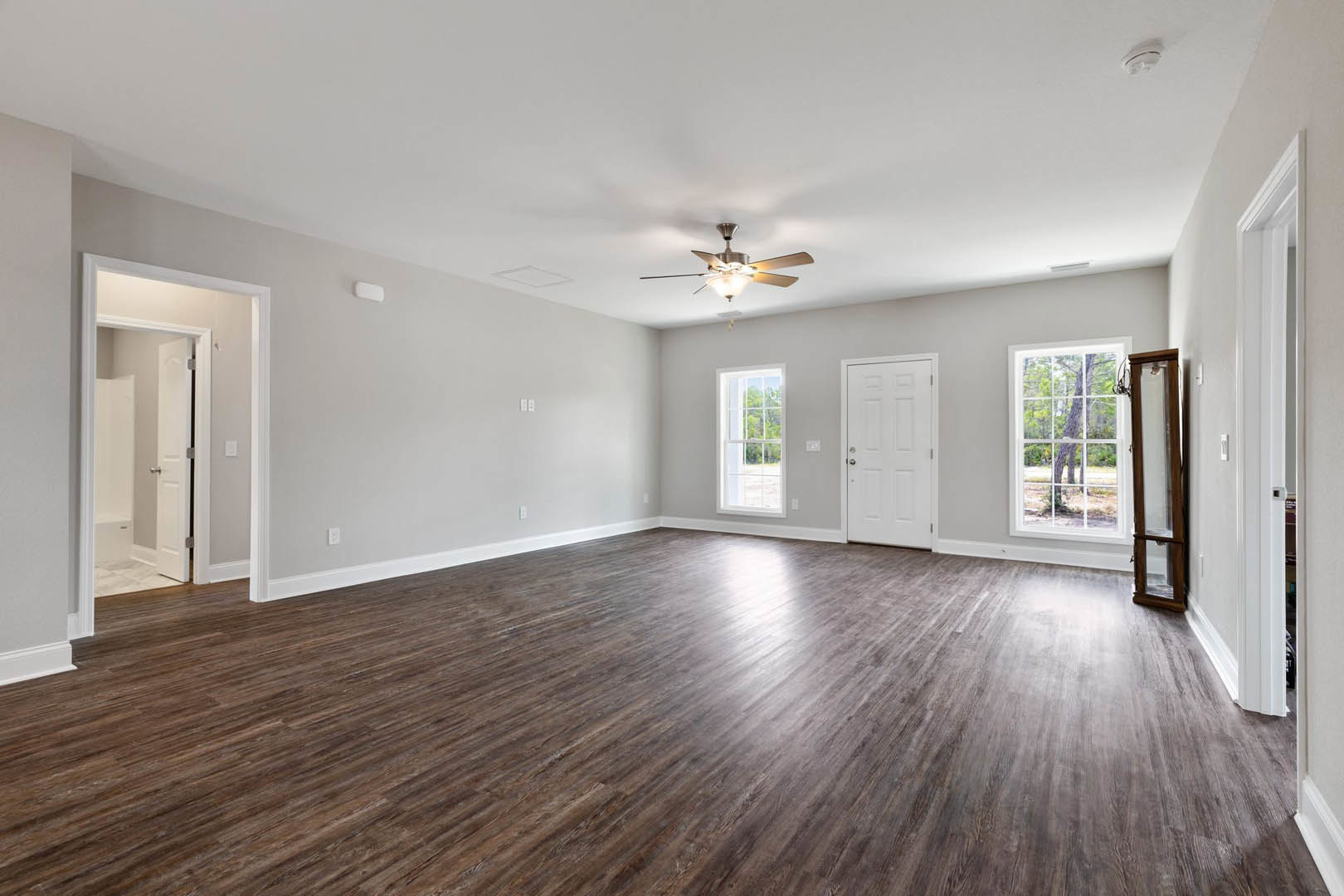Bright room with wood flooring, white walls, ceiling fan with light, large windows showing leafy tree, and white door with silver handle