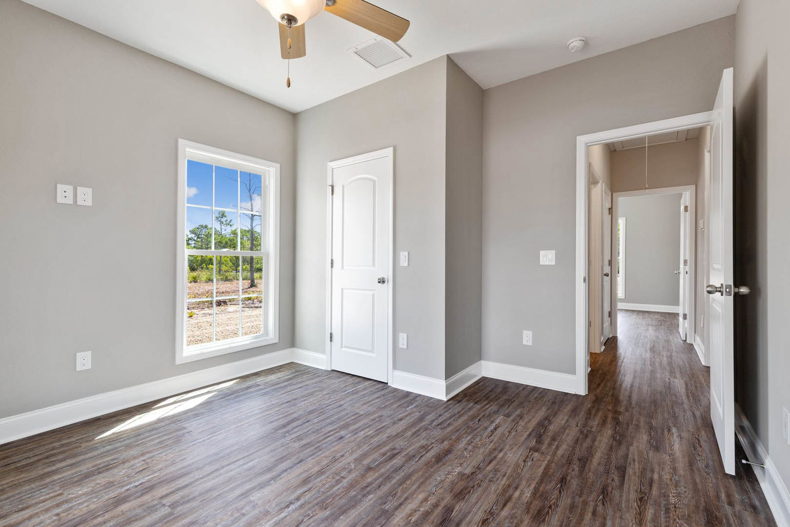 Wood flooring with white trim, ceiling fan, open white door with silver knobs, window overlooking trees and blue sky, ceiling vent