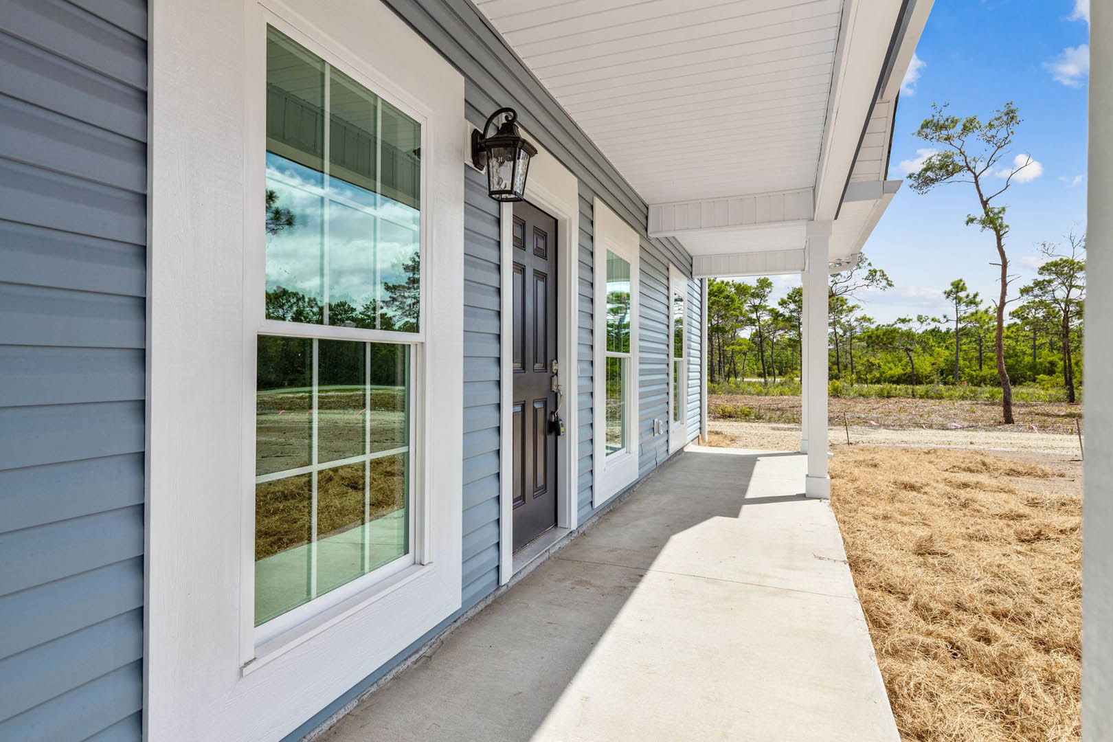 Wide front porch with white columns, hanging lantern, trimmed grass lawn, large windows reflecting trees and open field, slender tree branches visible near entry.