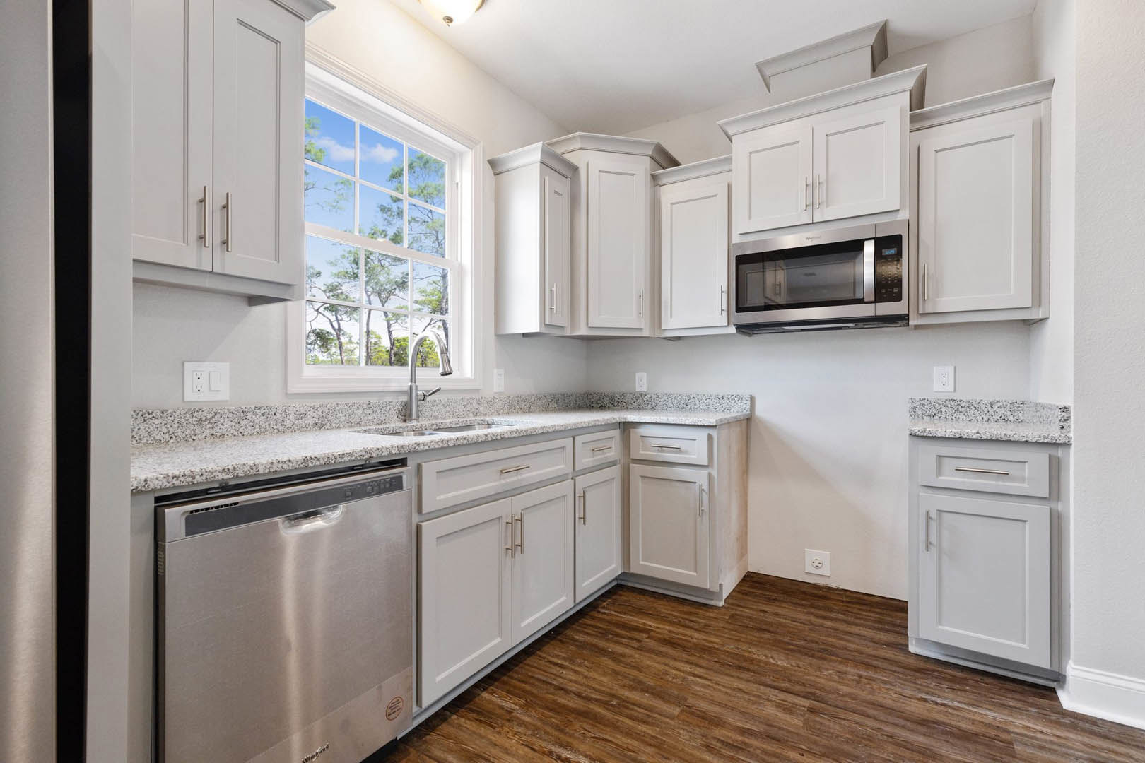 White kitchen cabinets, stainless steel dishwasher, glass door microwave, window with tree view, white outlet with black buttons, close-up of cabinetry.
