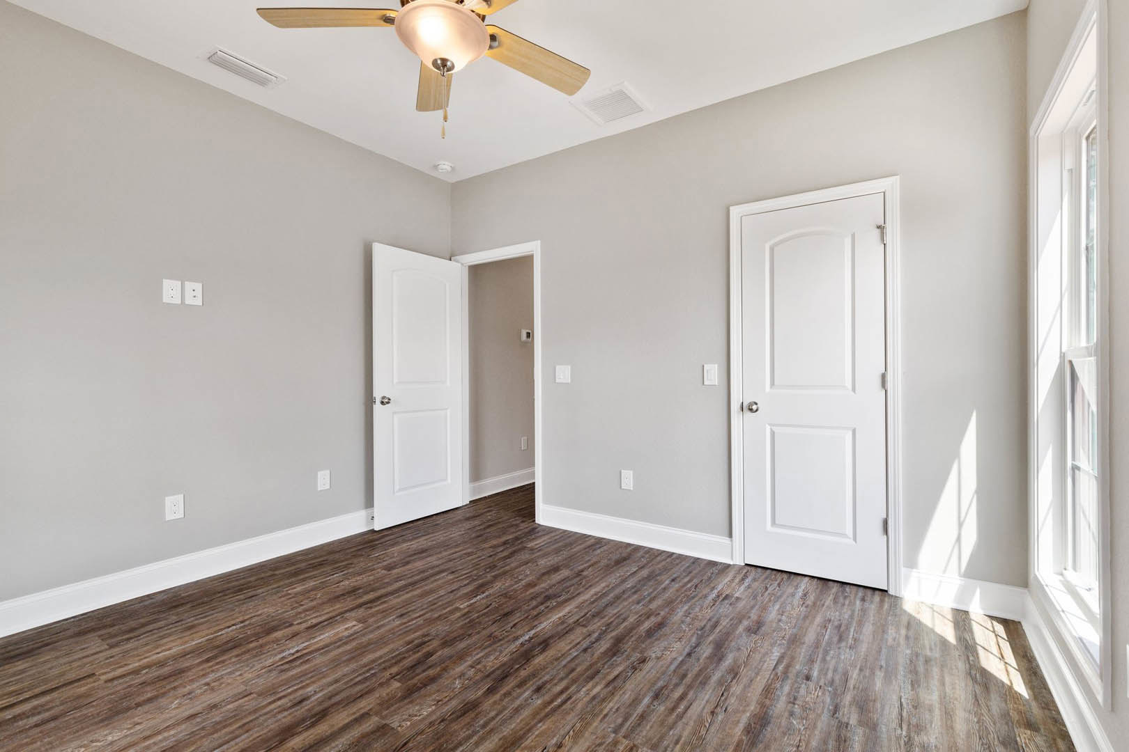 Wood flooring with white baseboards, two white doors with silver knobs, ceiling fan with light fixture, window with white trim
