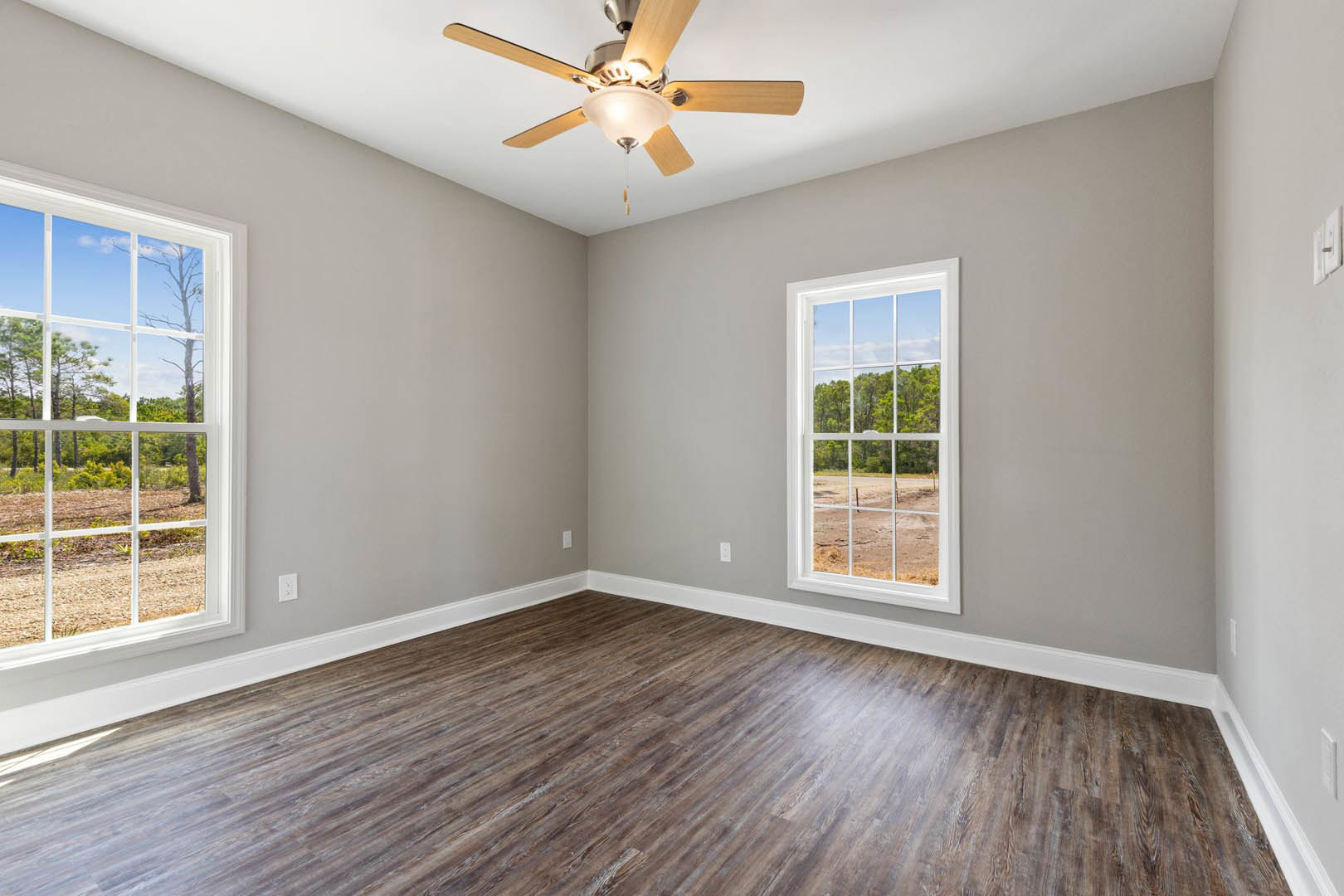Ceiling fan with light fixture above wood flooring, white trim, and window overlooking dirt road and trees