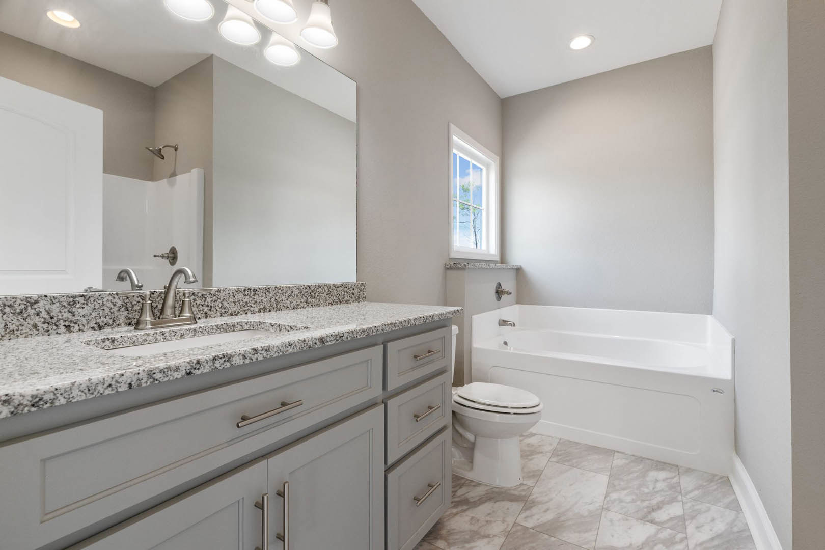 Bathroom with white tile floor, white bathtub beside toilet, rectangular sink with black faucet, window showing blue sky and clouds, light-colored cabinetry and countertop.