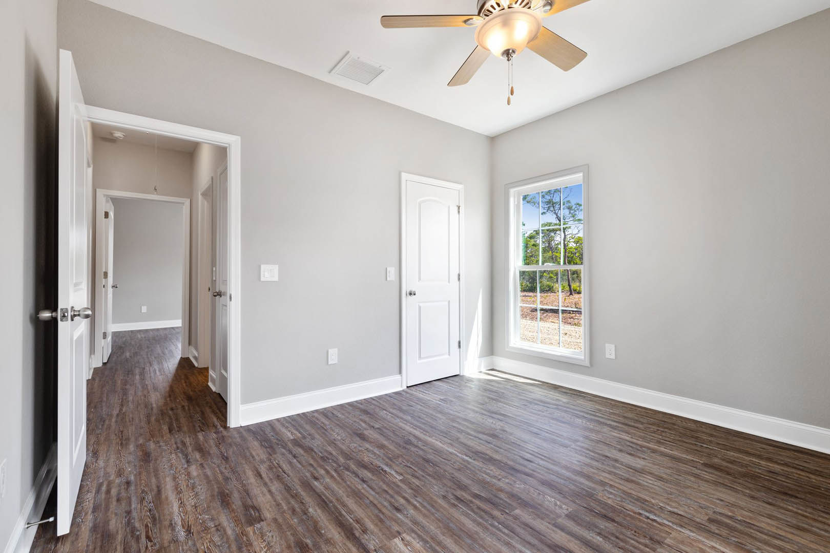 Wood flooring, white door with silver handle, ceiling fan with light fixture, window overlooking trees, neutral walls