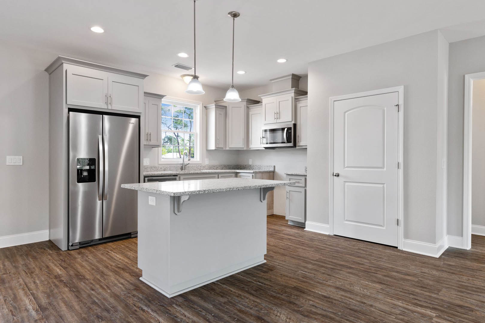 White kitchen with marble-topped island, stainless refrigerator, open microwave, white cabinetry with silver knobs, large window overlooking trees, light wood flooring