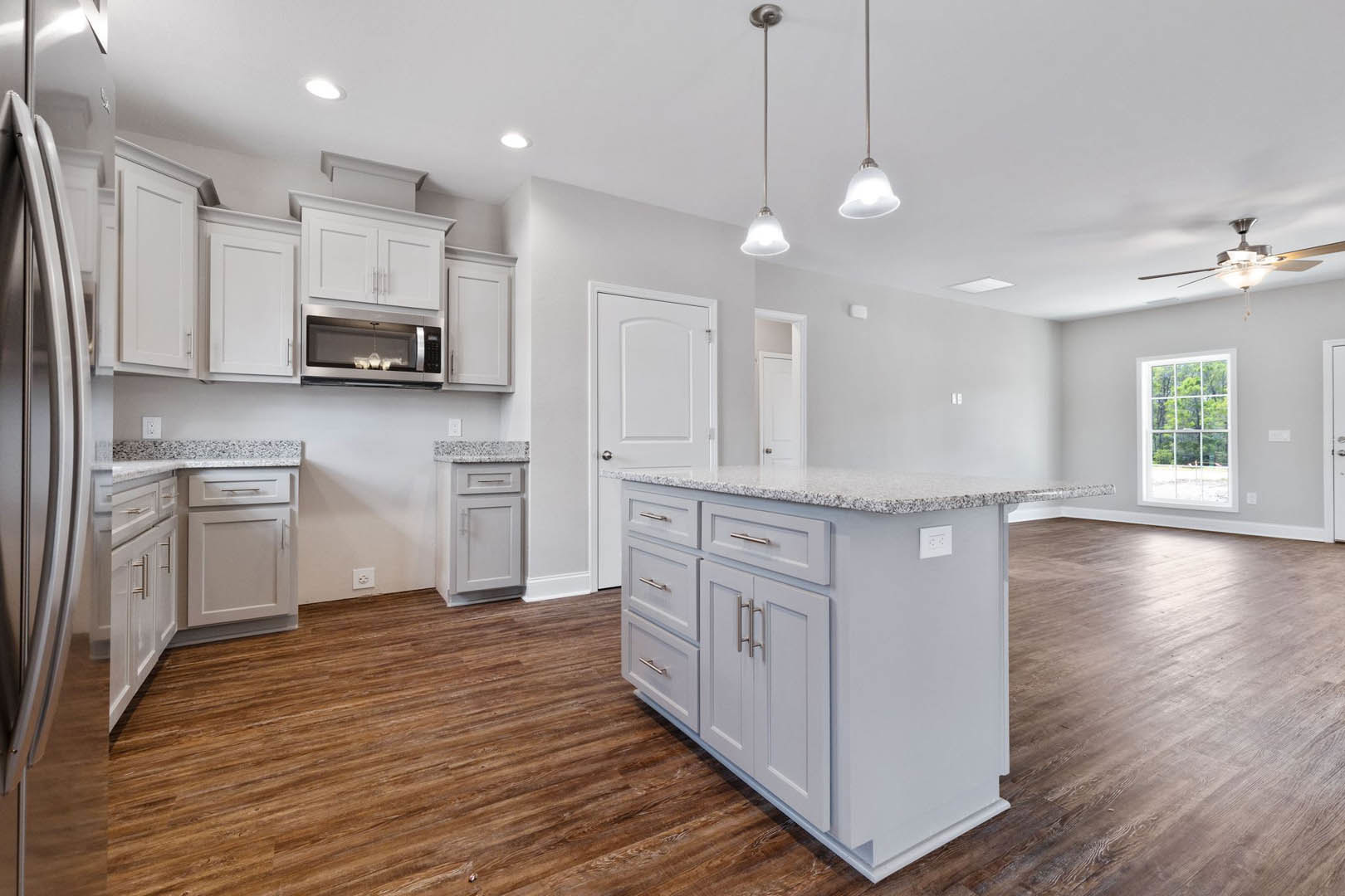 Kitchen with wide-plank wood flooring, white shaker cabinets, stainless steel refrigerator, multi-pane window, pendant light fixture, and stone countertops