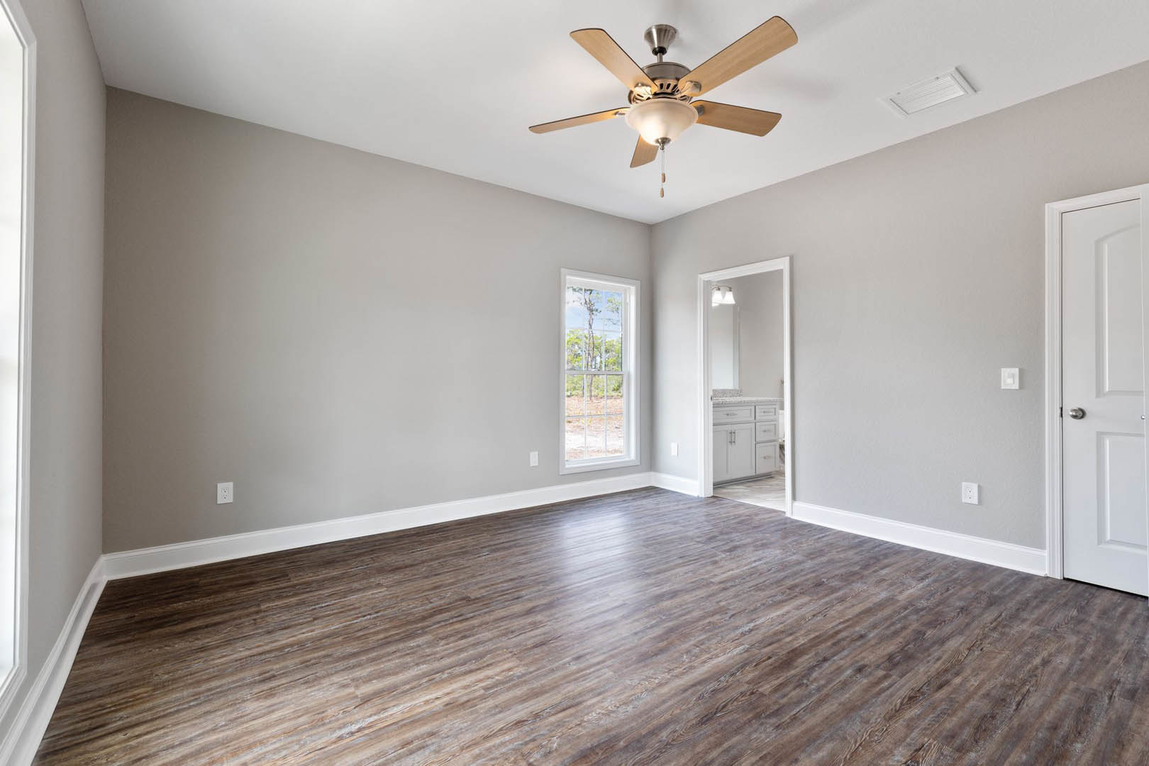 Ceiling fan with light fixture above hardwood floor, white walls, window showing trees outside, white door, marble bathroom countertop visible