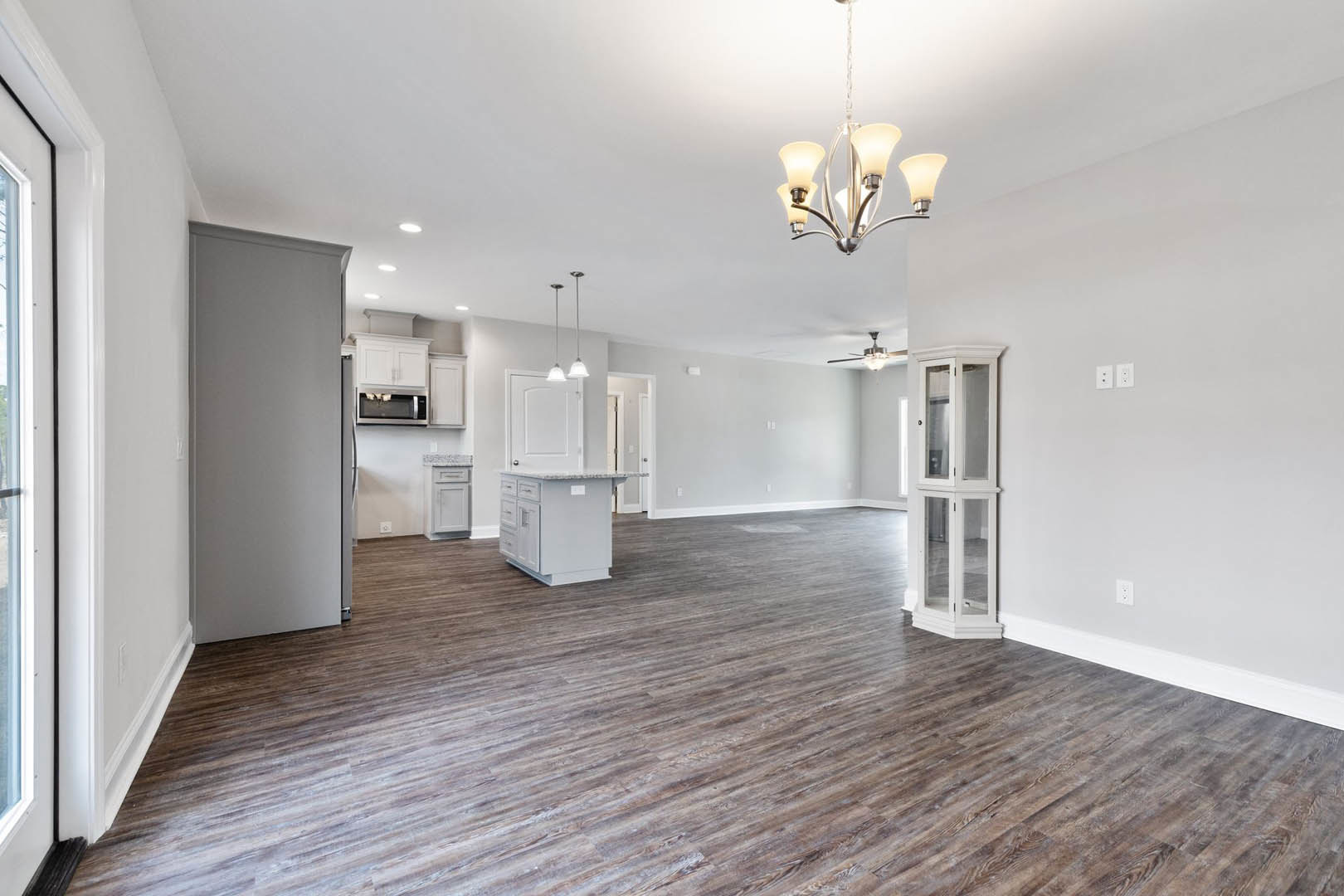 Spacious room with light wood flooring, white walls with black trim, ornate chandelier, adjacent kitchen area with matching wood floor, white cabinets with glass doors, built-in