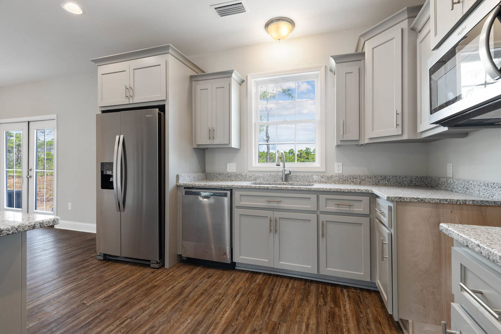 White kitchen cabinets, stainless steel refrigerator, built-in microwave, window above sink with view of trees, light countertops, wood flooring