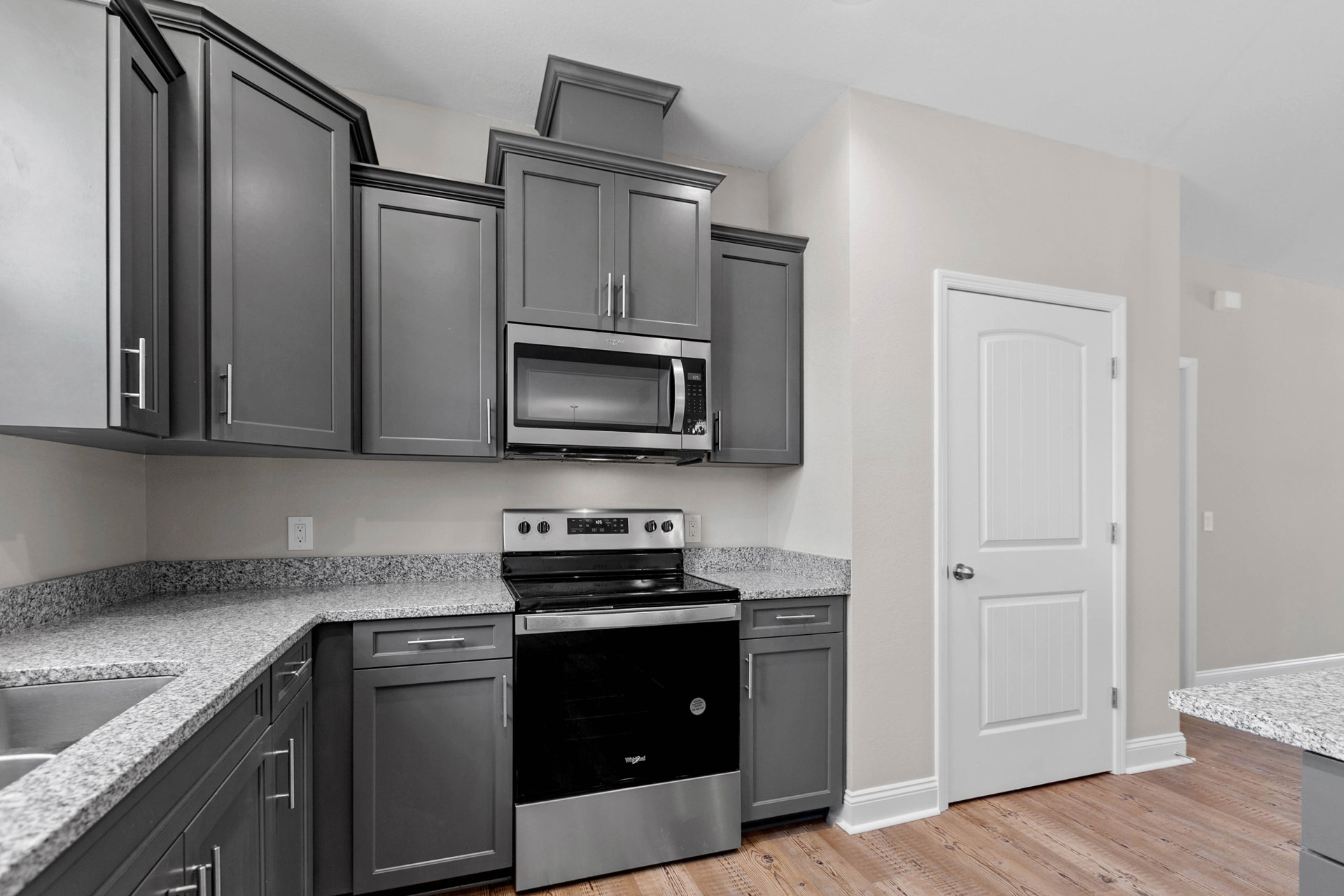 Grey kitchen cabinets with brushed silver handles, built-in microwave above a black and silver oven, white door with silver hardware, light stone countertop, and smooth grey