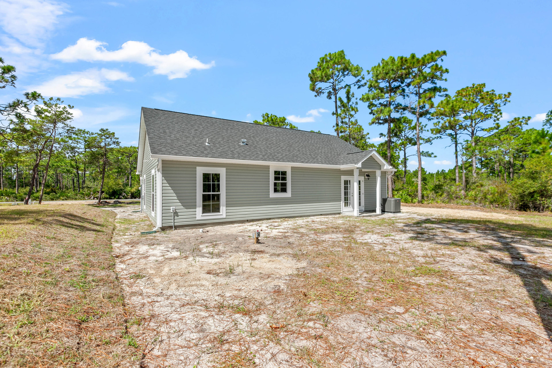 Two-story house with grey siding, white-framed windows, and a fire hydrant in the dirt front yard; mature trees and cloudy sky in the background.