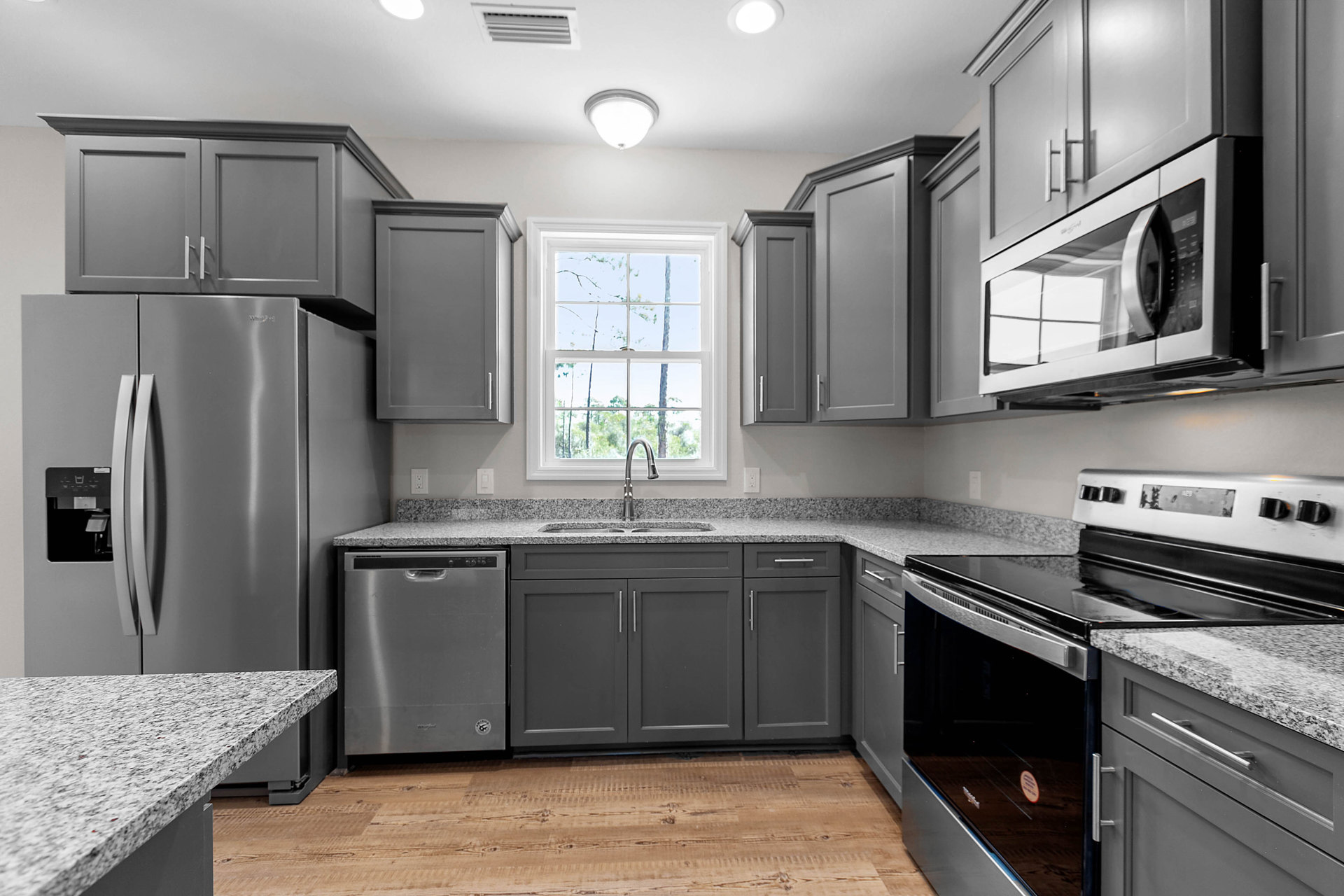 Grey shaker cabinets and black-and-white speckled granite countertops, stainless steel stove and oven, undermount sink beneath a window with trees visible outside, built-in