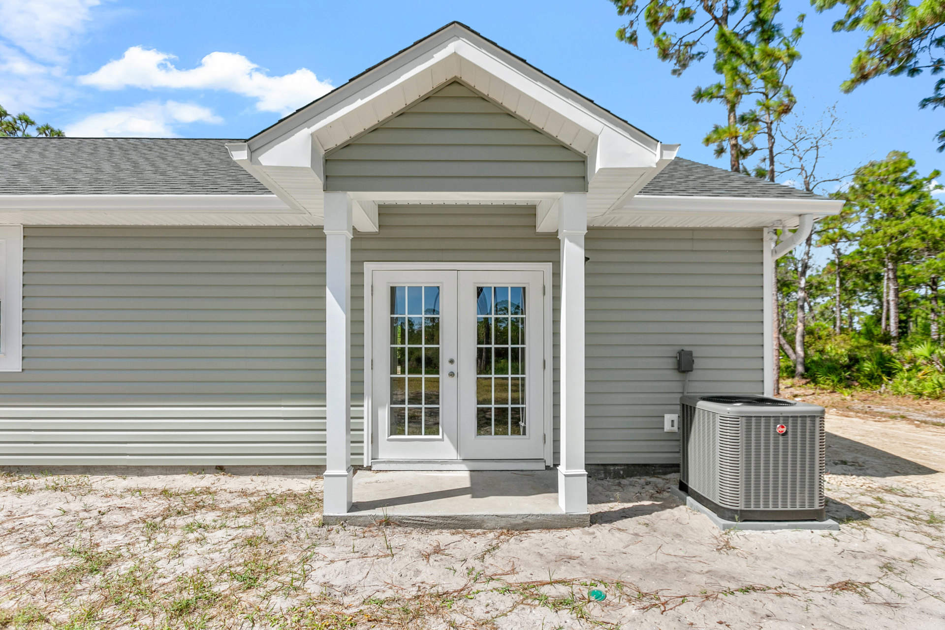 White double front door with glass panes, grey air conditioner unit beside entry, light-colored siding, small porch, tree branches visible in background
