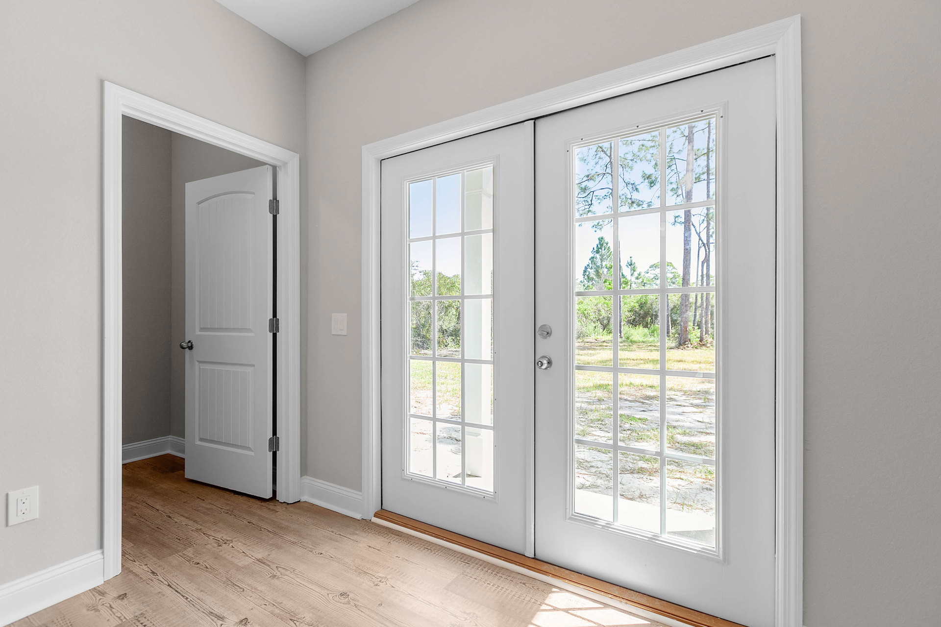 White double doors with glass panes and silver knobs set in a room with wood flooring, wall outlet, and a window showing trees outside