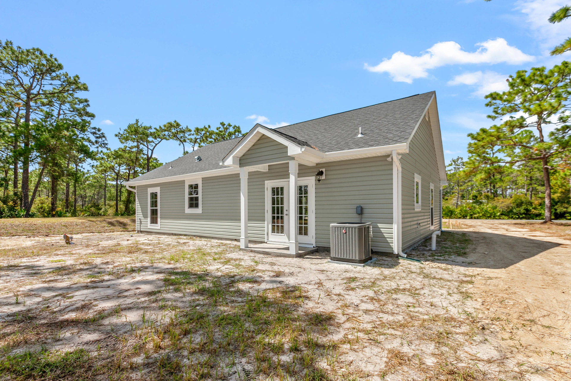 Two-story house with white door, heat pump unit beside foundation, surrounded by dirt yard and mature trees under blue sky