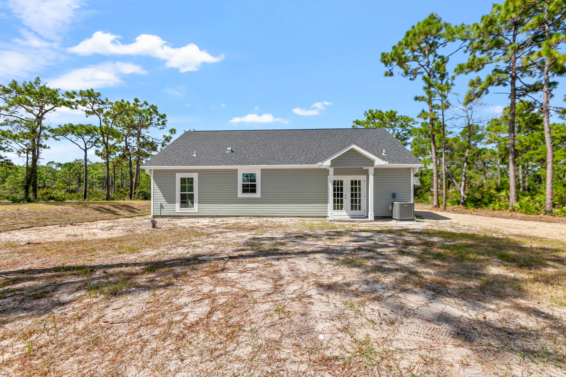 Two-story home with white-framed windows, glass double doors, light siding, grassy front yard, mature trees in background, and a grey air conditioner unit near the street.